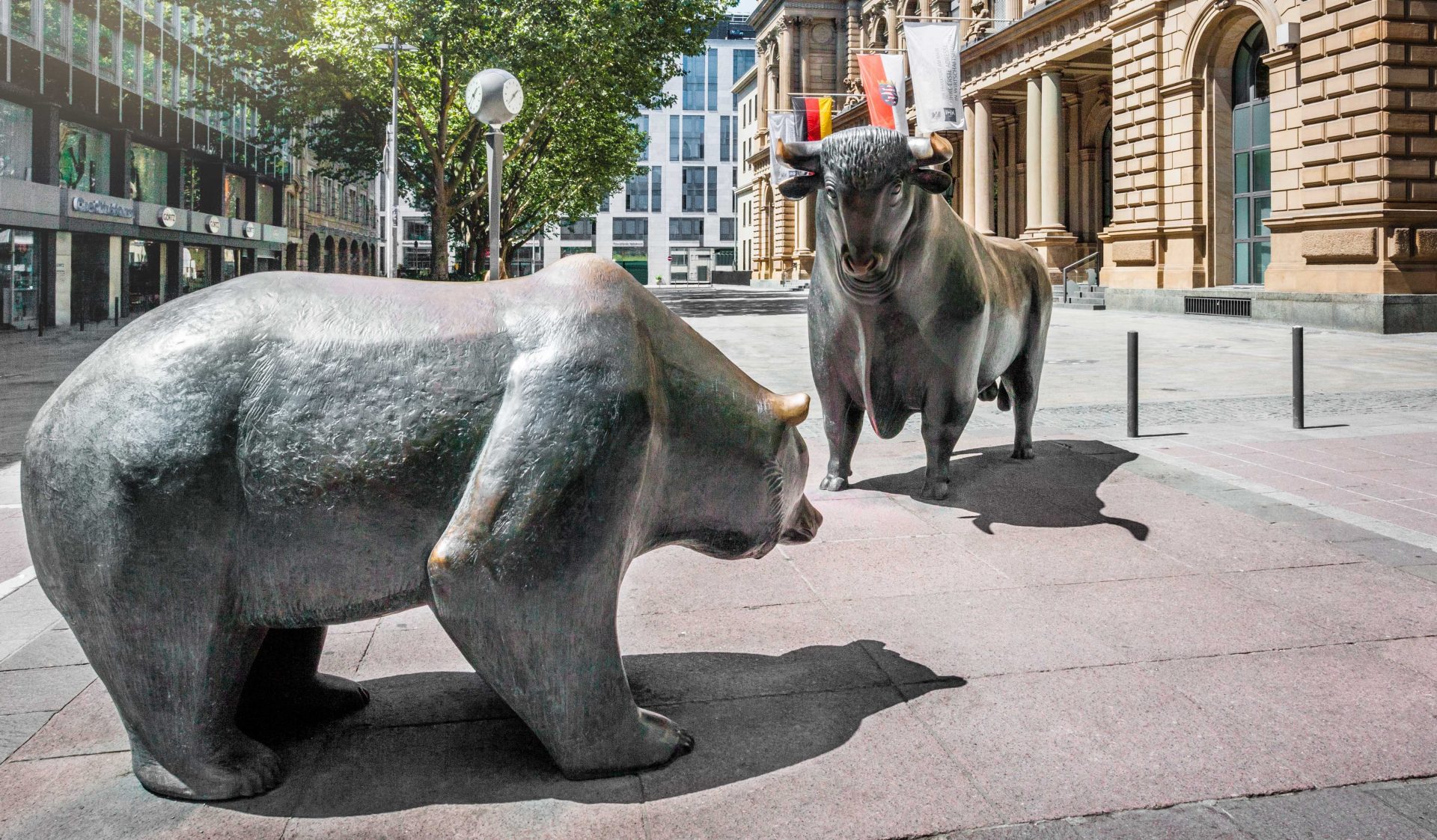 The image depicts two bronze sculptures of a bull and a bear facing each other on a street, likely representing financial market themes. The sculptures are situated in an urban environment with modern buildings in the backdrop and part of a historic building to the right. These figures symbolize the bull and bear markets in finance, where the bull represents a rising market and the bear signifies a declining market. The context suggests that this location could be near a financial district or stock exchange.
