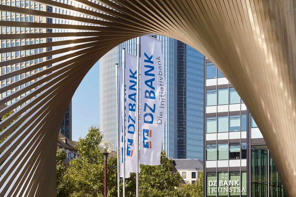 The image shows a modern architectural structure in the foreground, with buildings and skyscrapers in the background. There are flags and signage displaying "DZ BANK," indicating this is the location of a DZ BANK office. The structure in front creates an interesting arch framing the scene.