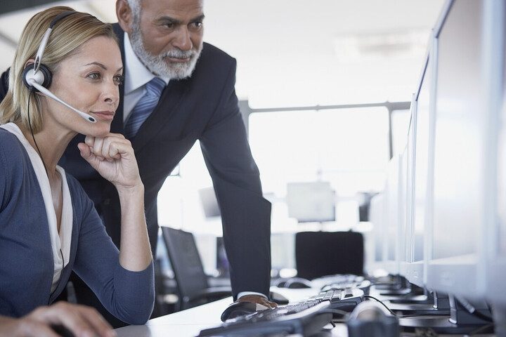 The image shows two people in an office environment. One person is sitting at a desk wearing a headset and looking at a computer screen, while the other person stands beside her, also looking at the screen. It appears to be a workplace or customer service setting, with multiple computer monitors and keyboards visible.