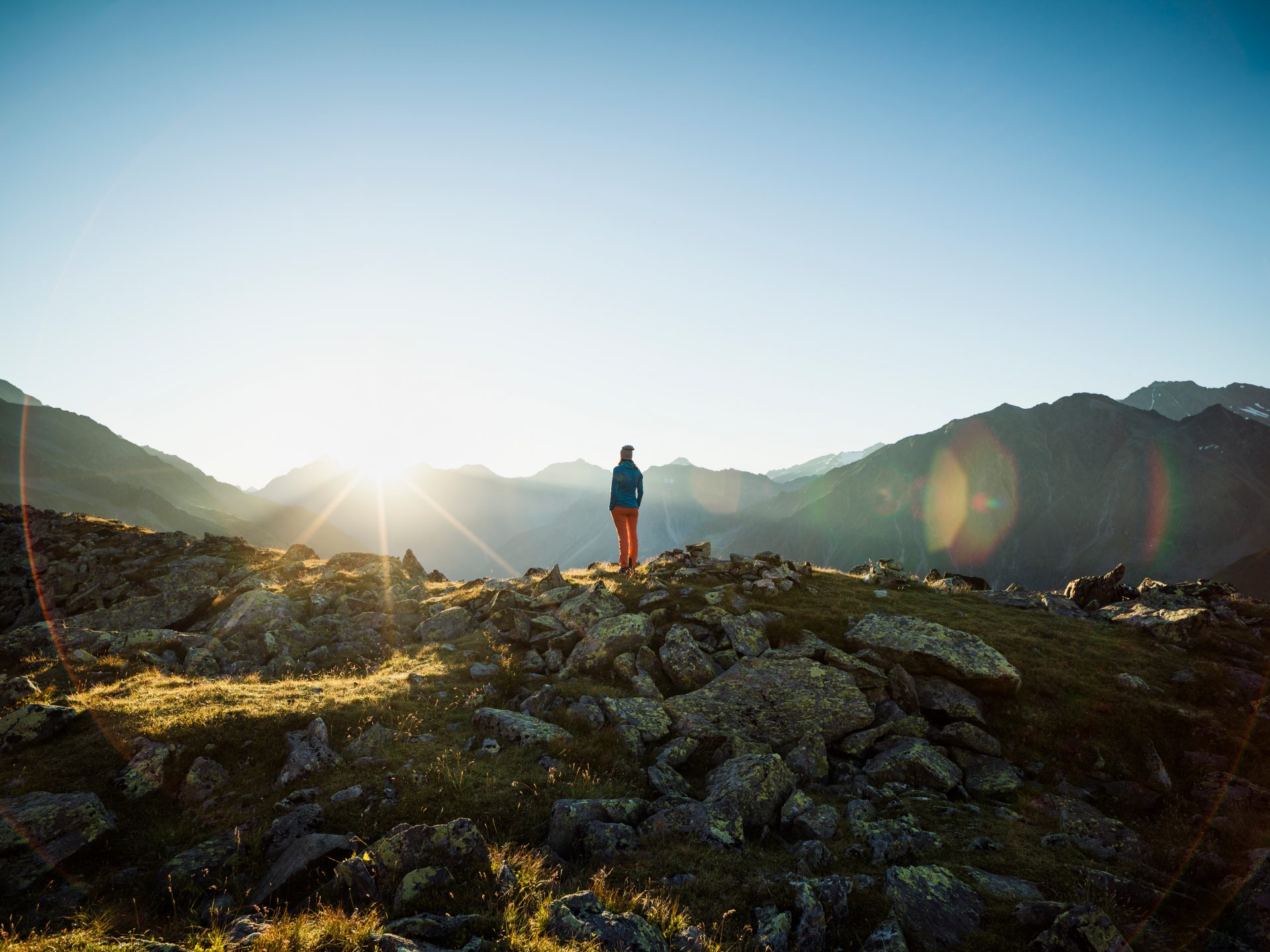 ### Description of the Image

**Scene:** 
- **Mountain Landscape:** The background reveals a beautiful mountainous setting with steep, rugged hills and peaks in the distance.
- **Sunrise or Sunset:** There's a prominent sun low in the sky, casting a bright light across the image, suggesting it's either early morning or late evening.

**Foreground:**
- **Person:** A lone figure is standing amid rocky terrain, facing the horizon where the sun is either rising or setting. The person is dressed in what seems to be outdoor attire suitable for hiking or trekking.
- **Rocks and Grass:** The terrain is rocky with patches of grass, indicative of a mountainous or highland area.

**Atmosphere:**
- **Lens Flare:** Noticeable lens flare effects suggest a direct exposure to sunlight, adding a dreamy or ethereal quality to the image.

**Overall Impression:**
- The image conveys a sense of solitude and contemplation, often sought by individuals exploring nature's vastness. The lighting hints at the beginning or end of a day's journey in the wilderness.