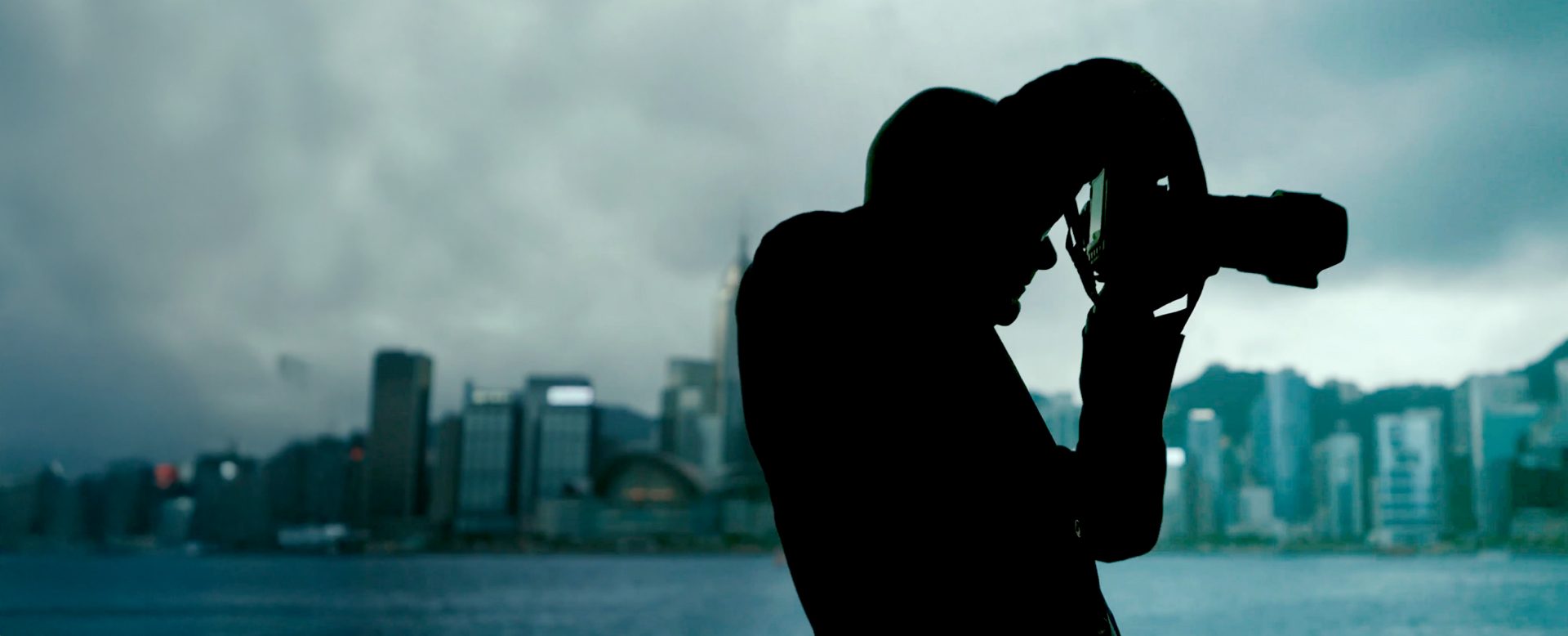 Silhouette of a person with a camera in front of Hong Kong’s skyline by the water; overcast sky.