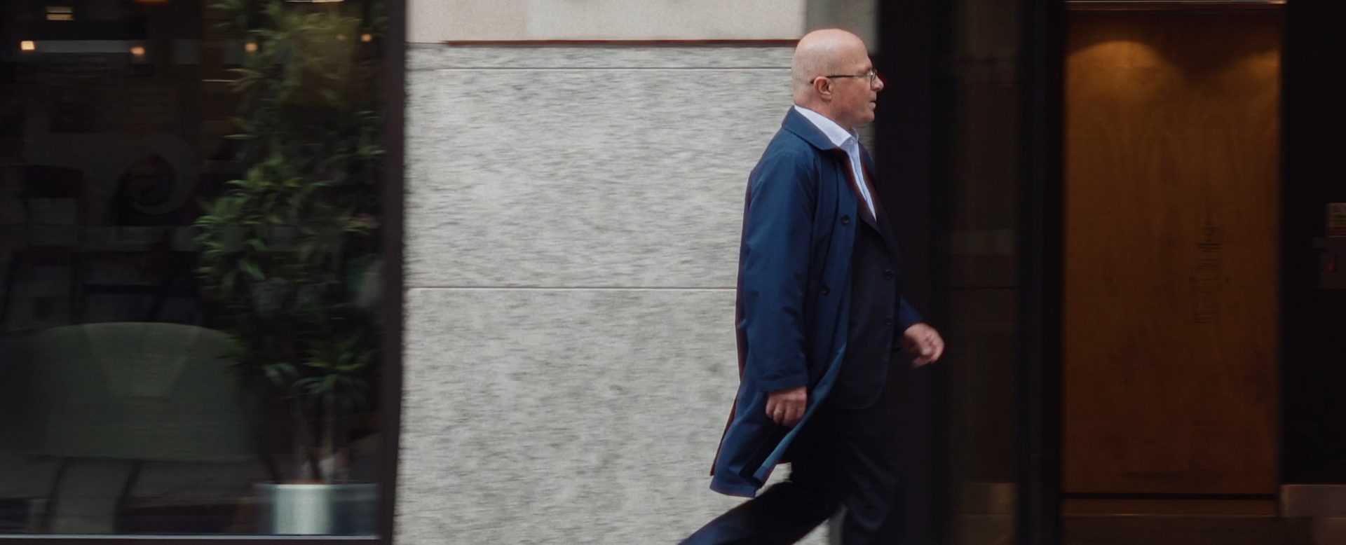 Person in a blue coat walking on the sidewalk past a building; entrance and plant visible through the window.