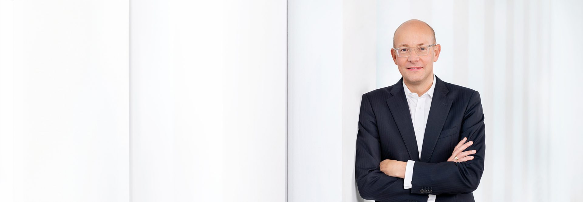 The image shows a person standing with arms crossed, wearing a suit and glasses, positioned against a light, minimalist background with vertical lines or columns. The overall setting appears professional.