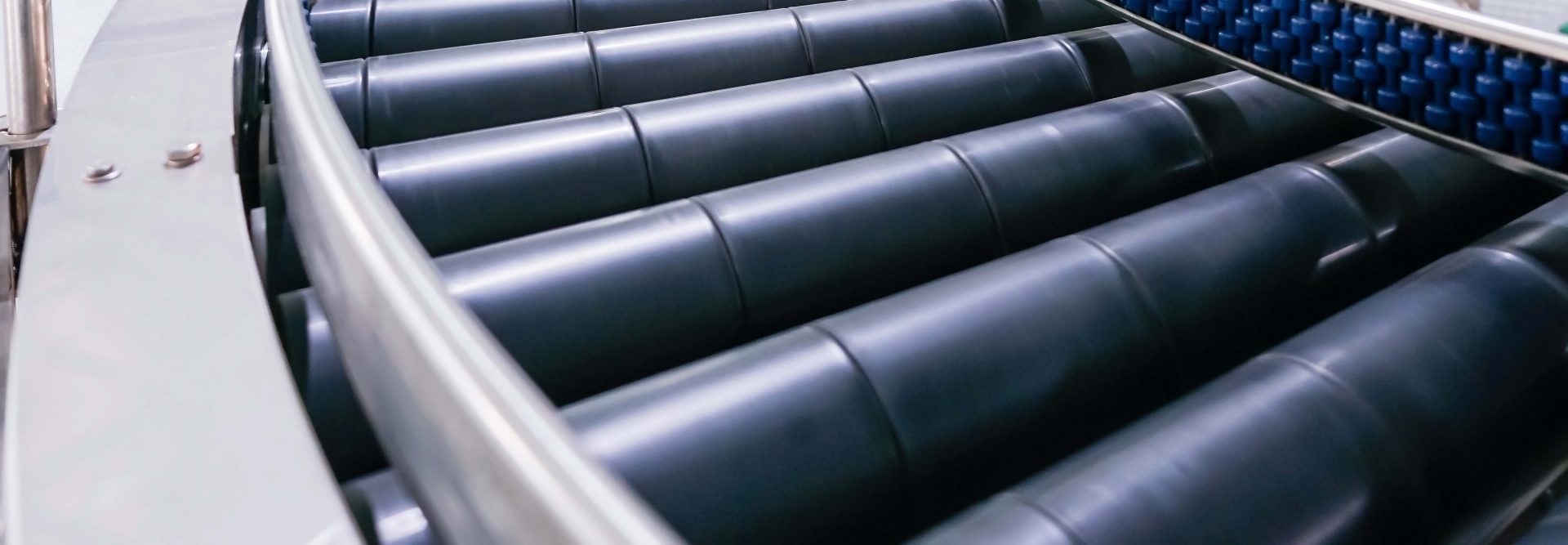 The image depicts a portion of a conveyor belt system. Details include:

1. **Conveyor Rollers**: 
   - Several cylindrical rollers form the moving part of the conveyor.
   - These rollers are designed to transport items through the system efficiently.

2. **Surrounding Frame**:
   - The conveyor is framed with metal or durable material that supports the rollers.
   - The frame provides stability and guides materials smoothly.

3. **Motion Mechanism**:
   - The image may show a section where transition occurs, moving objects from one section of the conveyor belt to another.
   - This can be witnessed in the incorporation of different roller surfaces or differing angles.

4. **Usage Context**:
   - Typically found in factories, warehouses, or similar industrial settings for transporting goods from one location to another.

5. **Machine Features**:
   - The structure might include additional elements like automated guiding or sensor systems to enhance operational efficiency.

This image focuses on capturing a close-up view of the mechanics that make up a conveyor system, highlighting industrial automation and product handling machinery.