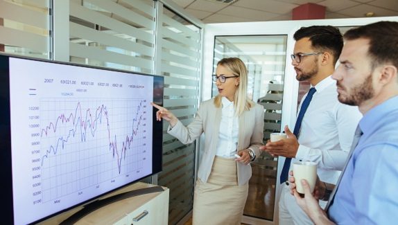 Presentation of a line graph showing data trends over time; people are gathered around the screen discussing the information.