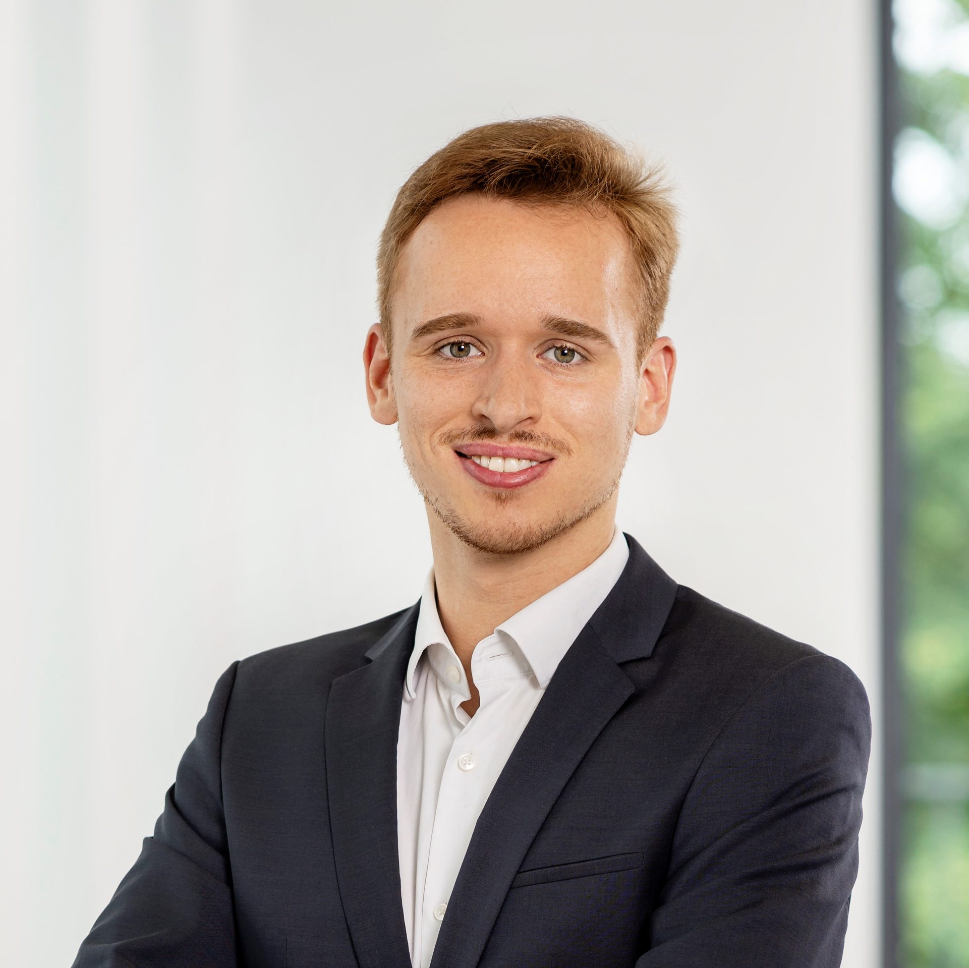 The image shows a person wearing a dark suit with a white shirt underneath. The background is blurred, likely indicating an indoor setting with a window or glass partition visible in the distance.