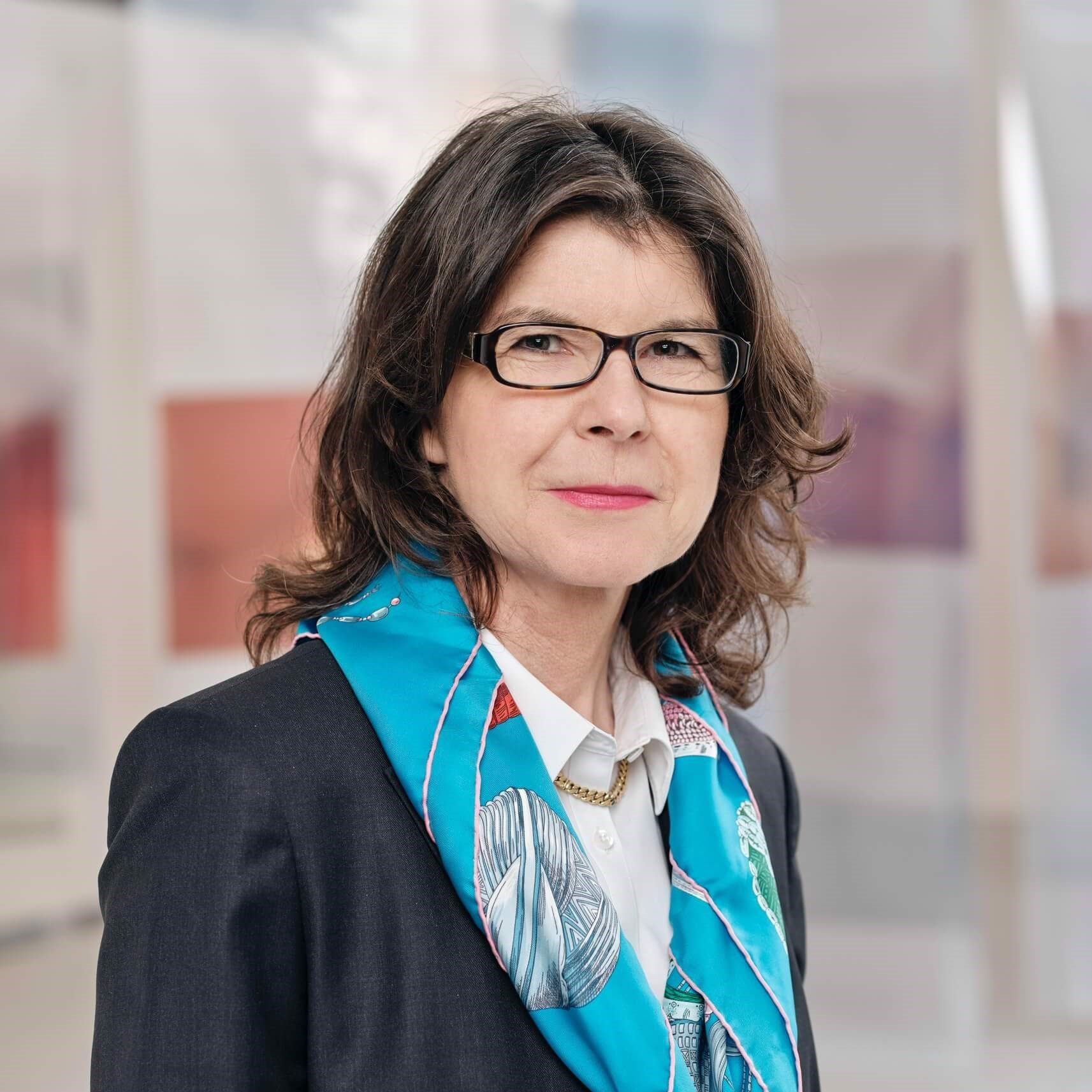 The image shows a person wearing a dark suit with a white shirt underneath. The suit is accessorized with a blue scarf that has patterns featuring colors such as grey, white, and light brownish-red tones. The background appears to consist of abstract or blurred elements in shades of beige and brown.