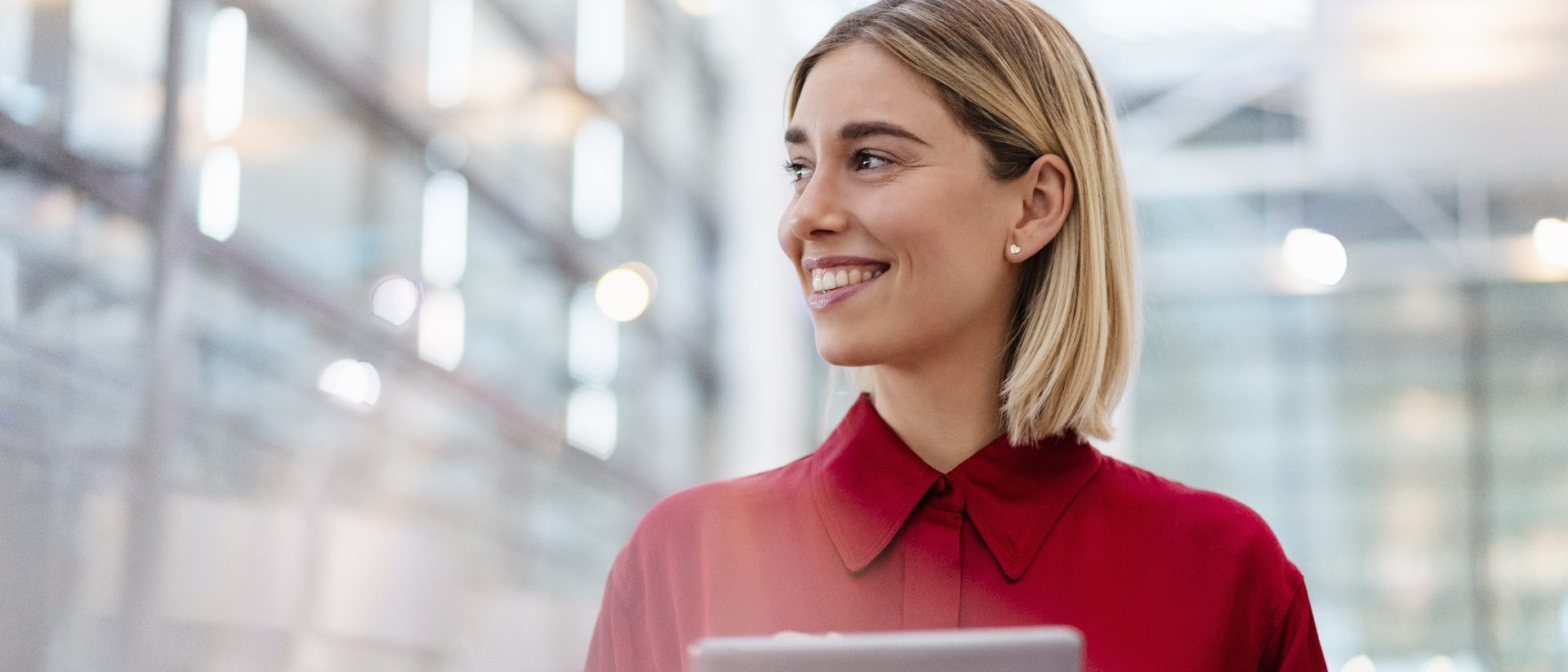 The image shows a person standing indoors holding a tablet in their hands. They are wearing a red shirt and are positioned in front of a bright and modern background with blurred lights and structures.