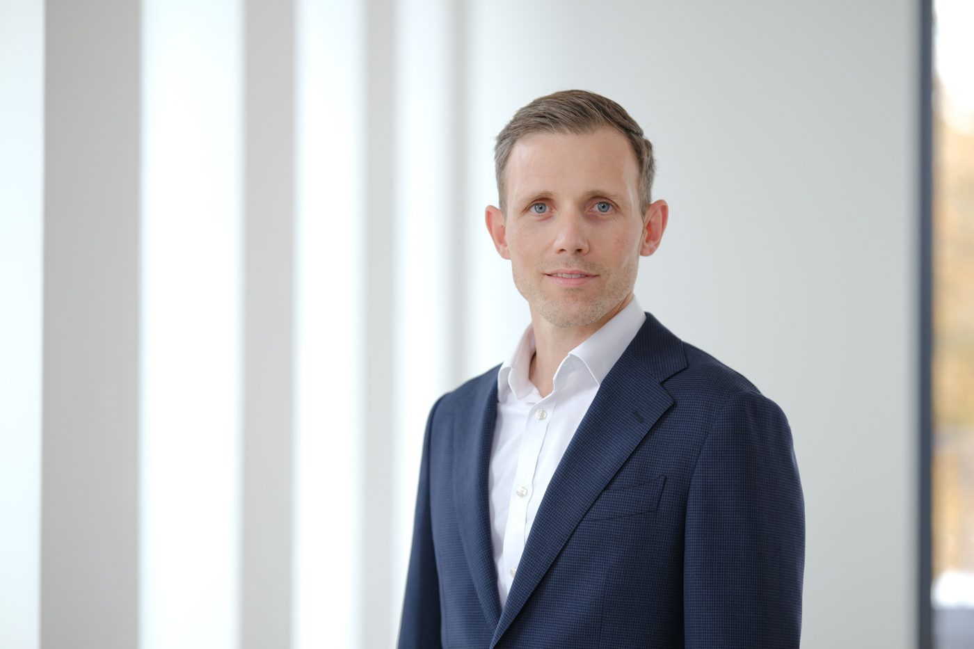 The image shows a person wearing a blue blazer and a white collared shirt in a bright indoor setting.