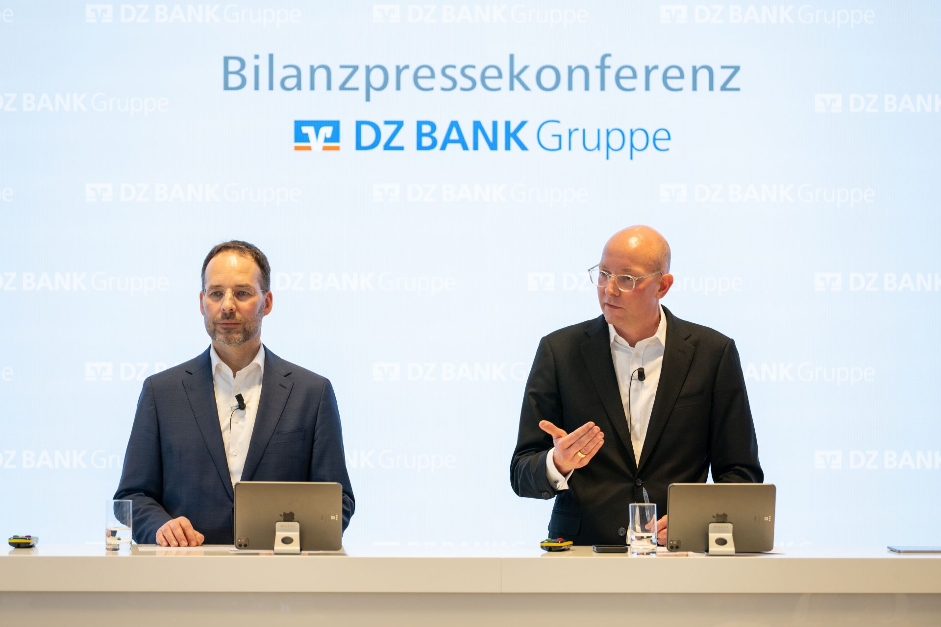 The image shows a press conference setup for DZ Bank Group, with two individuals standing at a podium with microphones and devices. The backdrop displays the text "Bilanzpressekonferenz DZ BANK Gruppe."