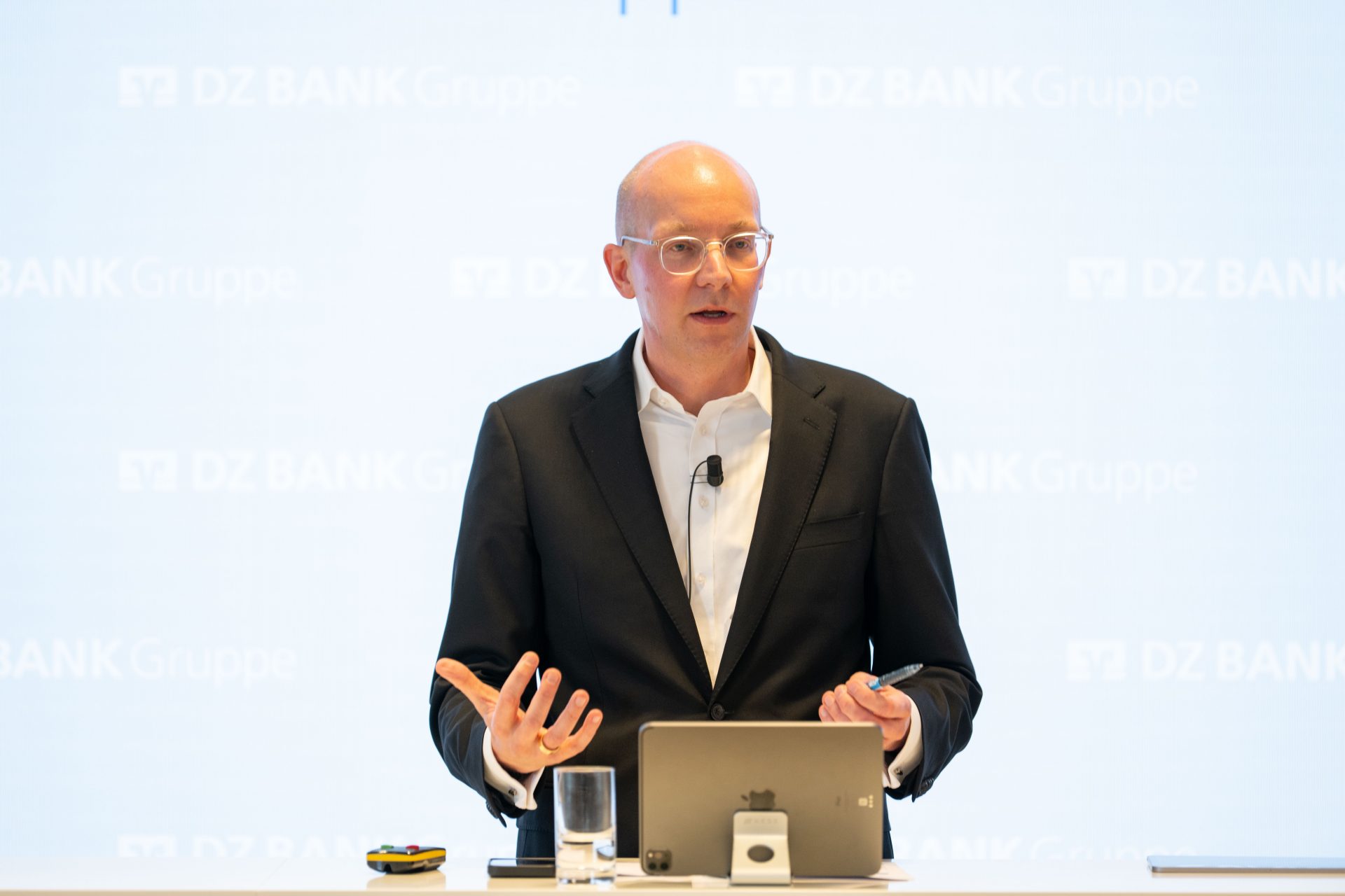The image shows a person standing at a podium with a microphone, a tablet, a glass of water, and a small electronic device or remote control. The background has "DZ BANK Gruppe" written on it, indicating a likely corporate or financial event.