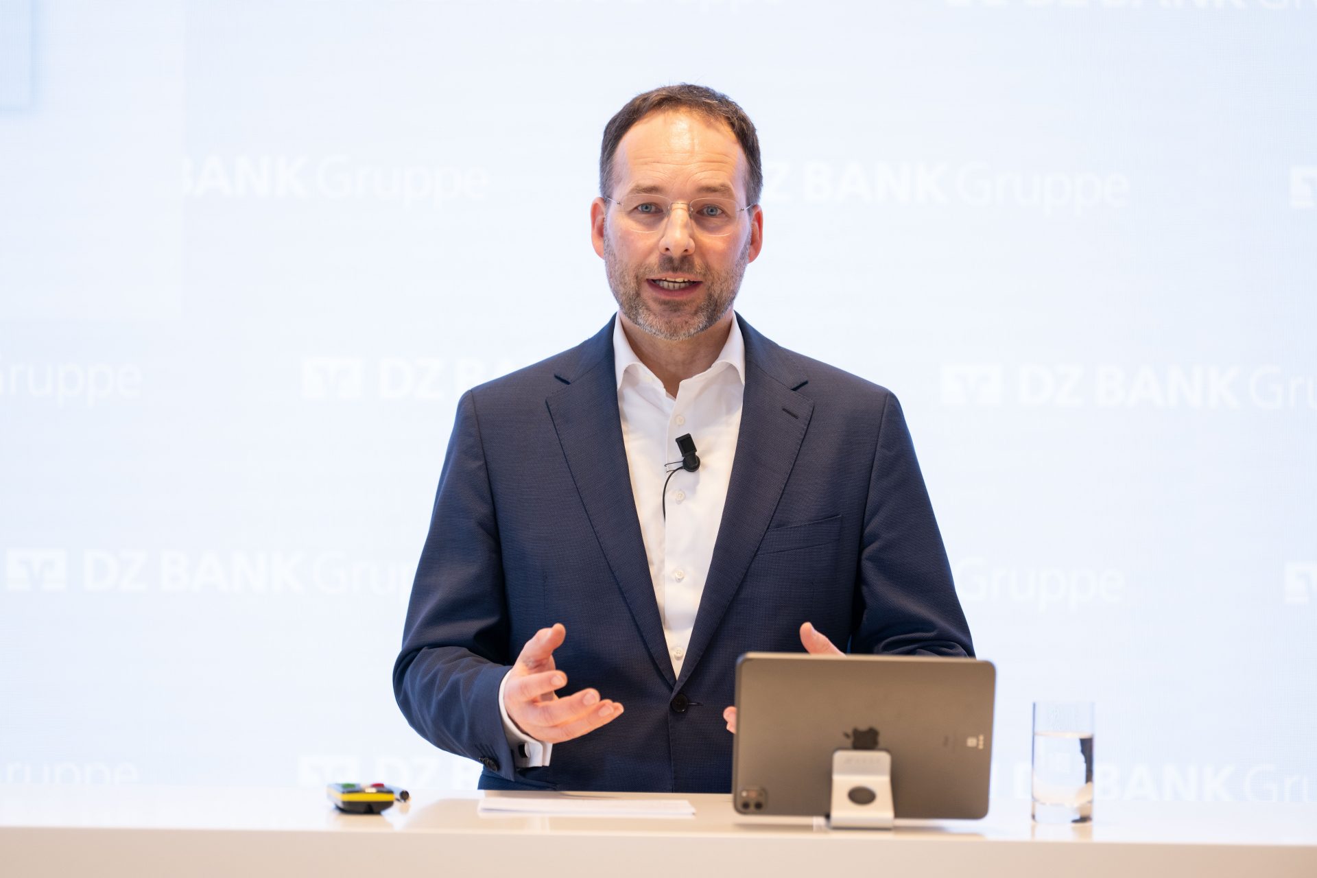 The image shows a person in a suit standing at a podium with a tablet and a glass of water, possibly giving a presentation or speech at an event associated with DZ BANK Gruppe.