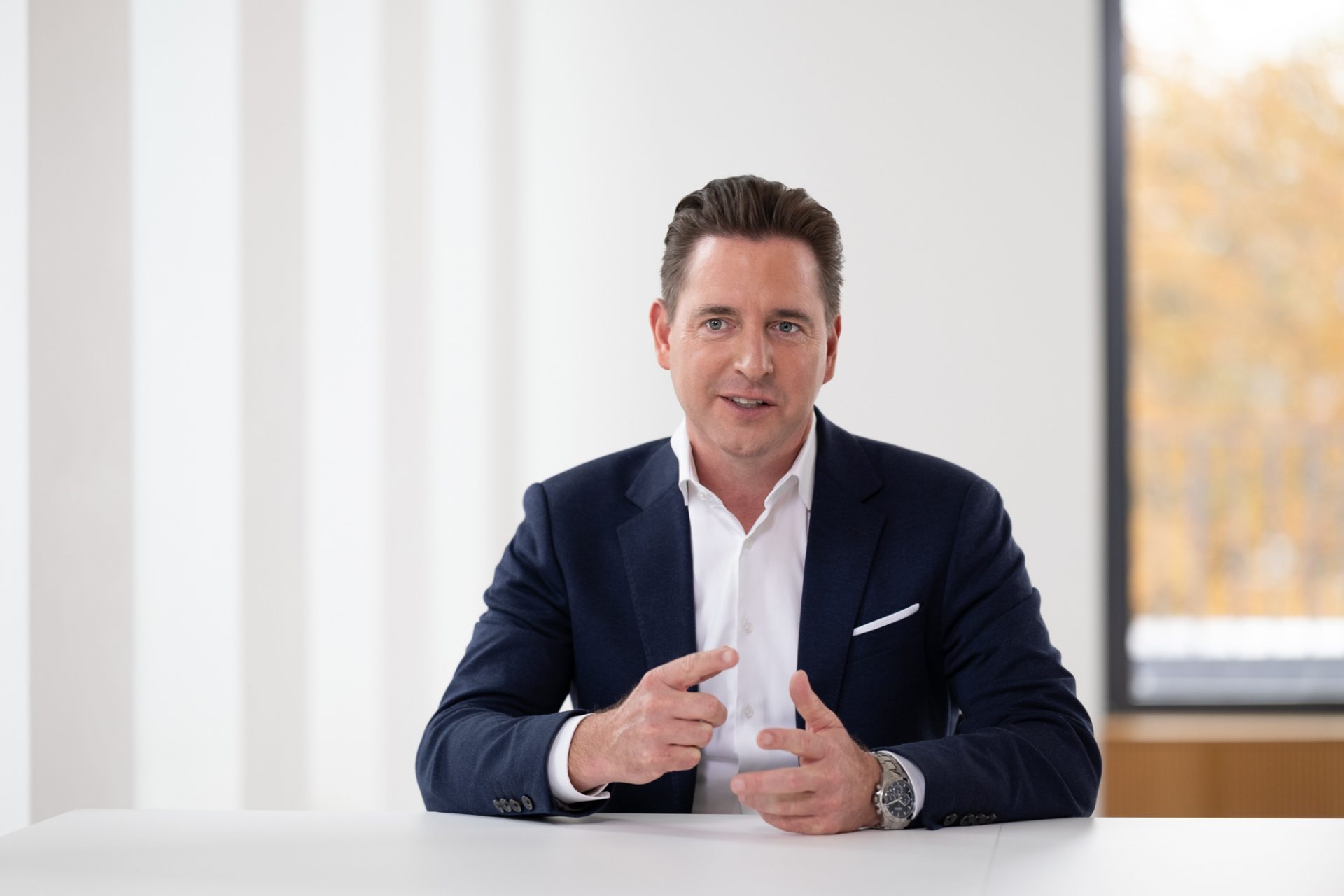 The image shows a person dressed in a formal suit, sitting at a table in a bright room with large windows. The view outside appears to be of trees or foliage in autumn colors.