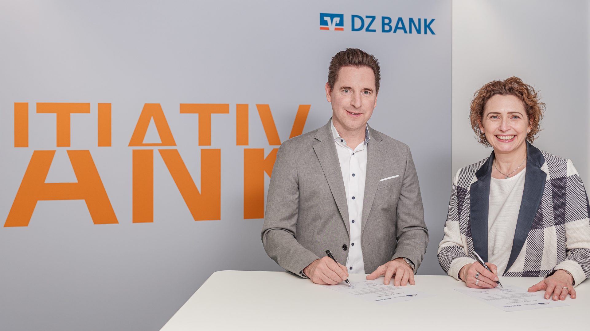 The image shows two people standing at a white table, appearing to sign documents. Behind them, there is a wall with partial large orange text and the logo of DZ BANK, a prominent German bank. It looks like a corporate or business setting, likely related to some official signing or agreement.