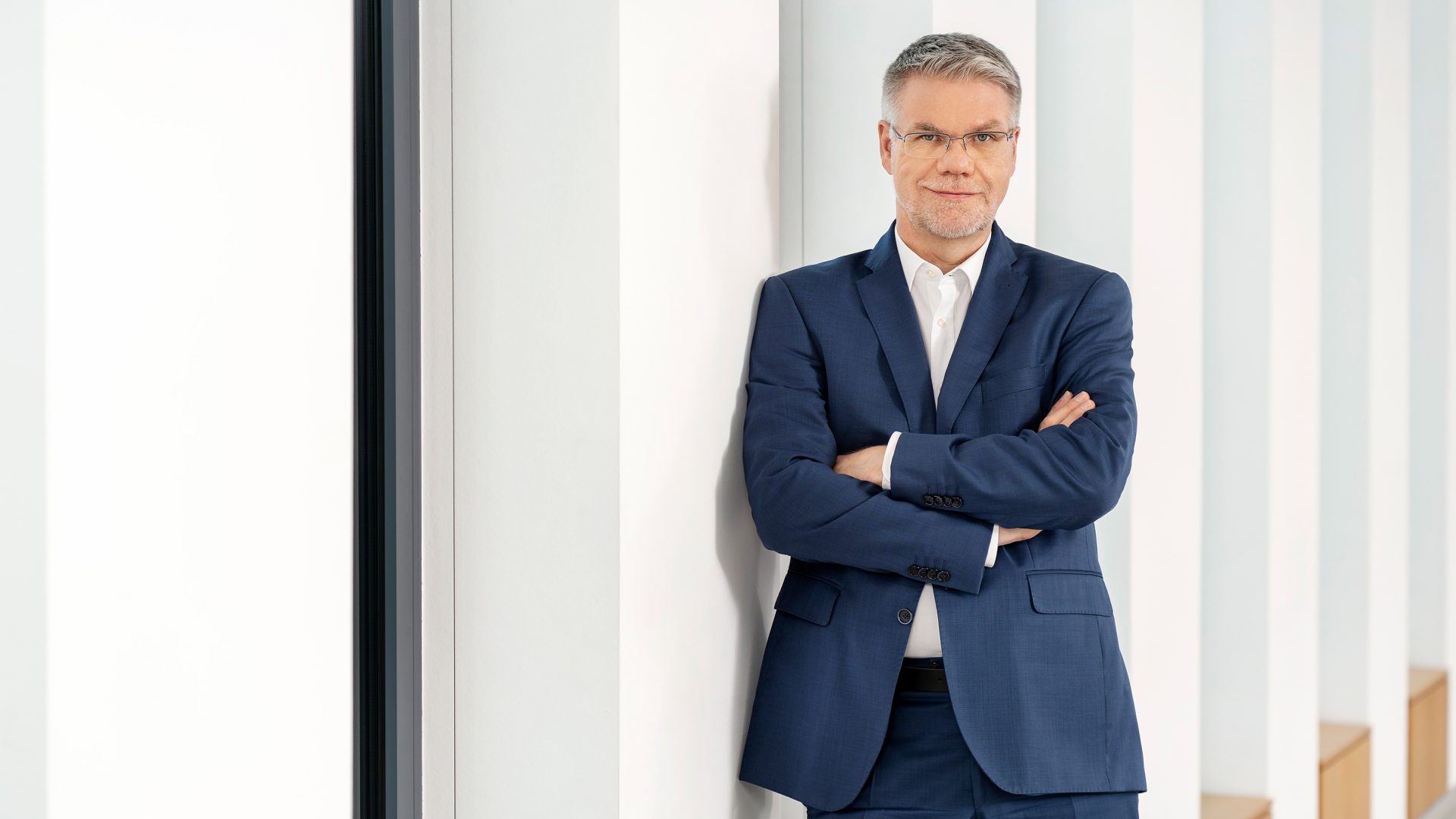 A man wearing a navy blue suit with a white shirt, standing against a wall with arms crossed.
