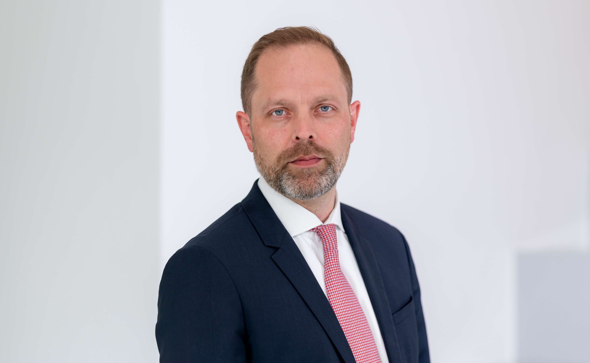 The image shows a person wearing a dark suit with a red and white striped tie, standing against a light, neutral background.
