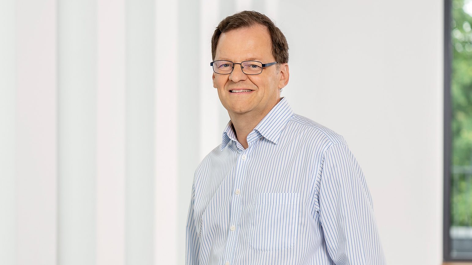 The image shows a person wearing a light blue striped shirt indoors, standing next to a white wall.