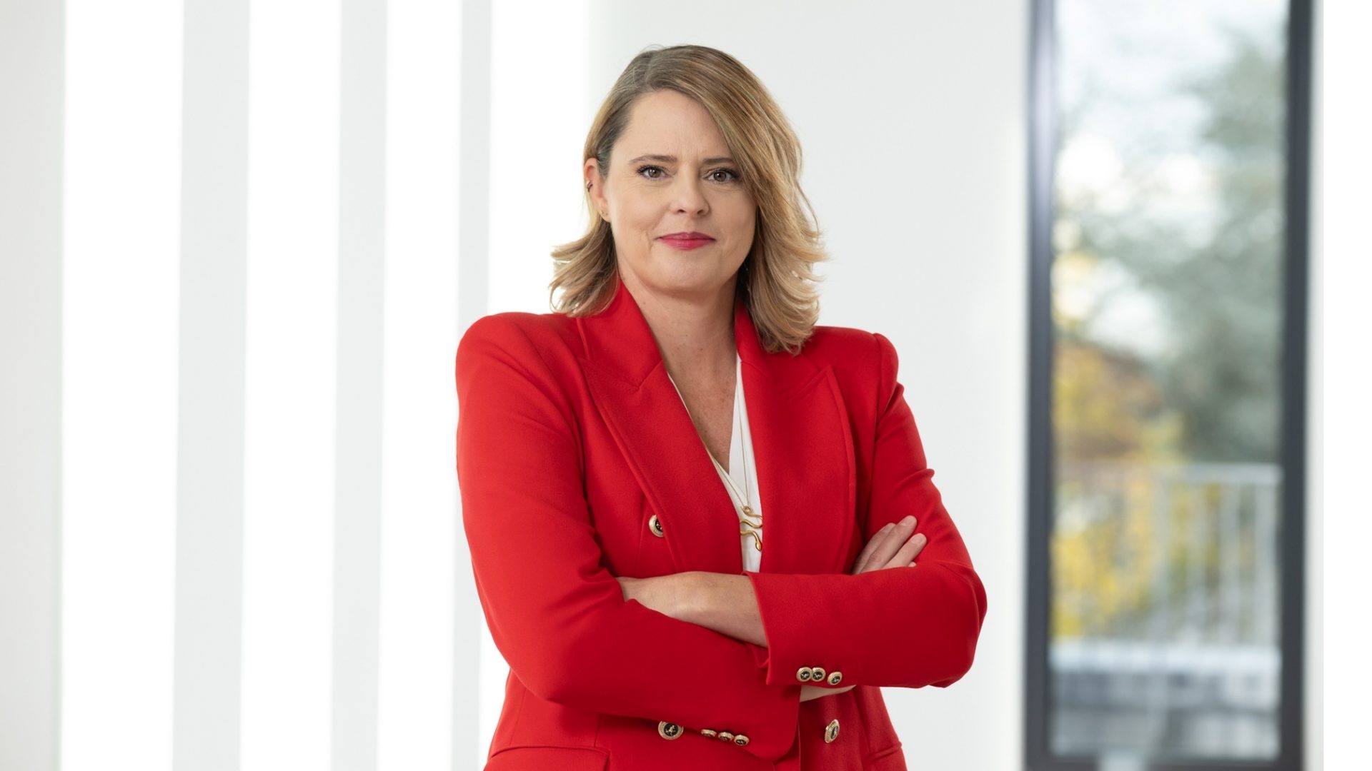A person wearing a red blazer with their arms crossed, standing indoors in a well-lit room with a window that shows part of the outside scene.