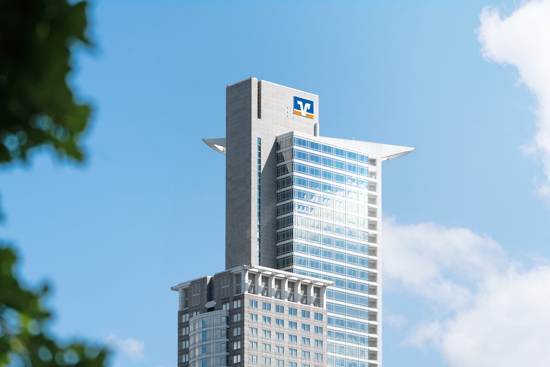 The image features a modern skyscraper in an urban environment. The building has a distinct architectural design with sleek glass windows and protruding sections at the top. It appears to be a commercial or corporate office building, given its size and styling.

**Details:**

1. **Building Design:**
   - **Structure:** The building is tall and has a rectangular form with additional structural elements extending outward, providing an artistic touch.
   - **Facade:** The facade is predominantly made of glass, reflecting the sky and surrounding light.
   - **Upper Features:** There are horizontal elements extending from the building's edges at the top, potentially for aesthetic or practical purposes.

2. **Environment:**
   - **Sky:** The sky is clear, with only a few clouds, suggesting a sunny day.
   - **Foreground Elements:** There are hints of green foliage in the lower corners, indicating trees or plants nearby.

3. **Logo or Signage:**
   - **Building Identification:** There's a logo or signage at the top that could identify the company or organization occupying the building, though it’s not legible in this description.

From this description, the image depicts a scenic view of a unique and potentially significant building in an urban area, captured on a clear day.