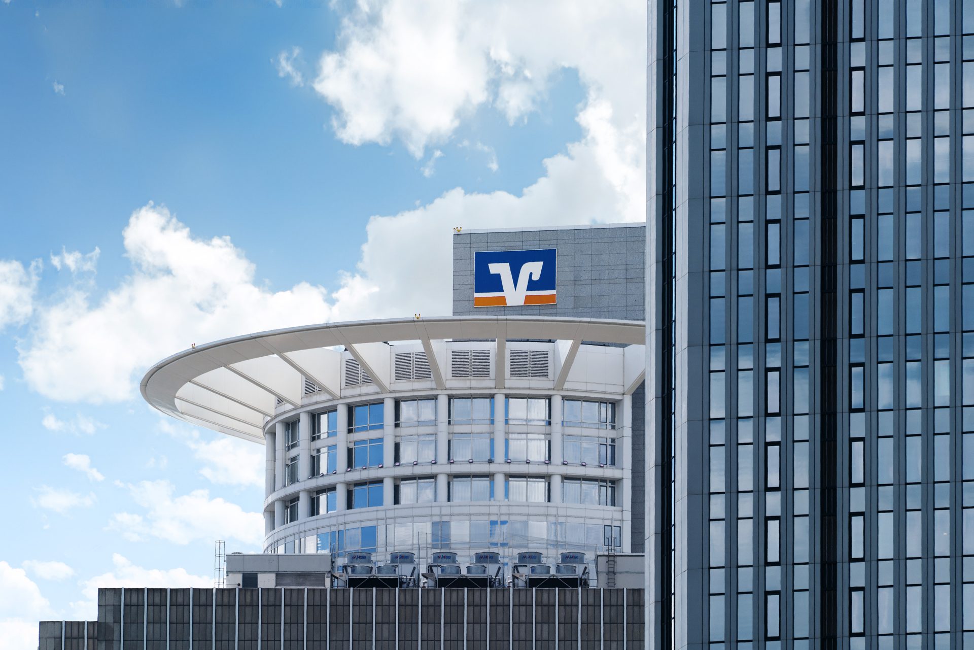 The image features a modern architectural building in an urban environment. The building has a distinct curved design on the top section, characterized by large glass windows and white framing. Prominently displayed on the roof is a logo that appears to be related to Volksbank or a similar financial institution, as indicated by the "V" symbol in the logo. The backdrop of the image includes a clear blue sky with scattered clouds, contributing to the image's bright and airy atmosphere. Also visible are the vertical lines and structures of adjacent buildings, highlighting the contemporary cityscape.