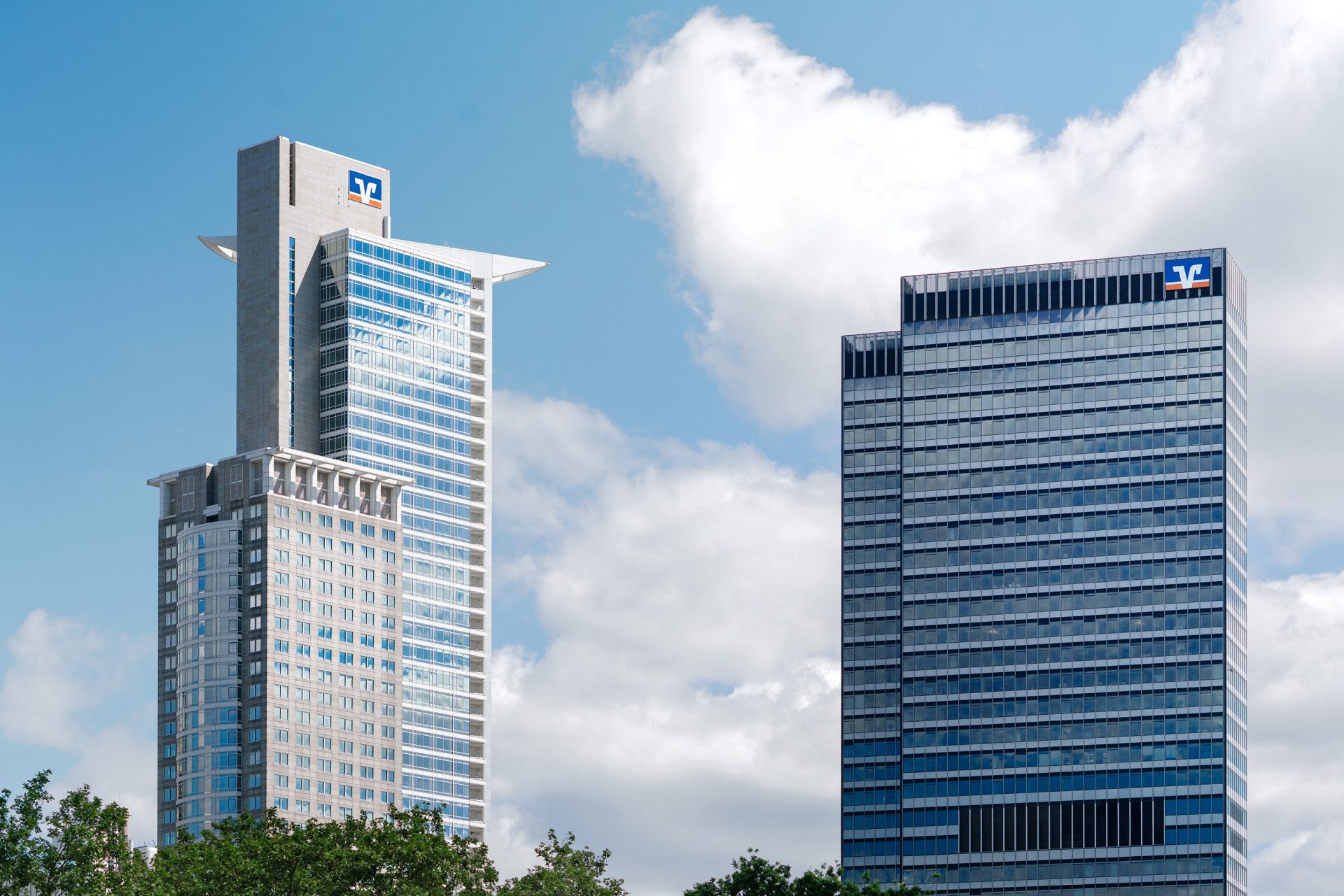 The image features two modern skyscrapers in an urban environment. They are positioned against a backdrop of a partly cloudy sky, offering a contrast between the architectural structures and the natural elements.

### Key Components:

1. **Skyscrapers**:
   - **Left Building**:
     - Taller and slimmer structure.
     - Features a sharp, angular design at the top that adds to its distinctive profile.
     - Windows are symmetrically placed, providing a sleek and streamlined appearance.
   - **Right Building**:
     - Wider and slightly shorter than the building on the left.
     - Uniform, horizontal rows of windows give it a more traditional rectangular shape.
     - Both buildings appear to be commercial or corporate establishments.

2. **Brand Logo**:
   - Both buildings display a logo at the top, indicating possible corporate headquarters or offices.
   - The logo appears to be affiliated with a finance or banking institution.
 
3. **Natural Elements**:
   - **Sky**:
     - Bright blue with patches of fluffy, white clouds, suggesting a clear day.
   - **Greenery**:
     - Trees and foliage are visible at the bottom of the image, hinting at a well-planned urban landscape that incorporates natural elements.

### Contextual Analysis:
- The architectural style suggests a focus on modernity and functionality.
- The presence of logos atop both buildings denotes a commercial zone, likely in a financial or business district.

### Visual Appeal:
- The juxtaposition of architectural precision with the softness of nature creates an aesthetically pleasing urban scene.

To summarize, the image captures the essence of urban sophistication balanced with natural beauty in a bustling city environment.