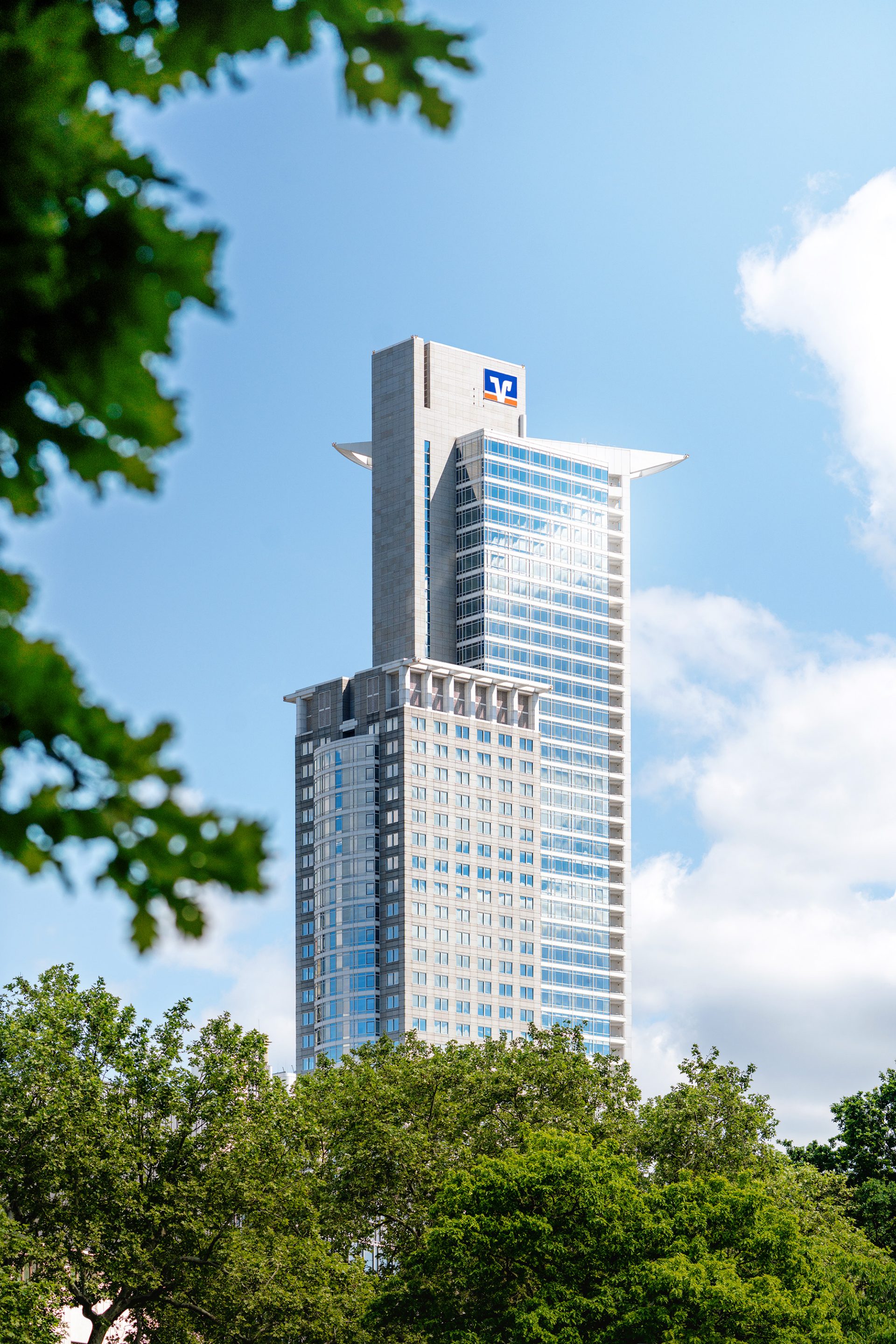 ### General Image Content

1. **Modern Skyscraper:**
    - The image prominently features a modern skyscraper with numerous glass windows.
    - The design is sleek with both vertical and horizontal elements.
    - It appears to be a tall, multi-story building.

2. **Tower Composition:**
    - The building consists of a main tower structure with smaller auxiliary structures.
    - Architectural features include a protruding section or overhang near the top.

3. **Logo or Branding:**
    - There is a logo or insignia near the top of the building indicating possible corporate or brand presence.

4. **Background Environment:**
    - Greenery and trees are visible in the foreground and to the side, suggesting an urban environment with landscaped areas.
    - The sky is mostly clear with a few clouds, indicating a bright day with good weather.

5. **Angle and Perspective:**
    - The photograph is taken from a ground-level perspective, looking up at the skyscraper, capturing its grandeur.

6. **Context and Setting:**
    - This type of building is commonly found in developed cities and business districts, serving mixed uses such as office space, residential apartments, or hotel accommodations.

### Identifying the Building
Given the lack of explicit identifiers other than the logo and architectural style, further information or location details would be needed to accurately identify the skyscraper. For example, the logo might provide hints regarding the building's purpose or owner.