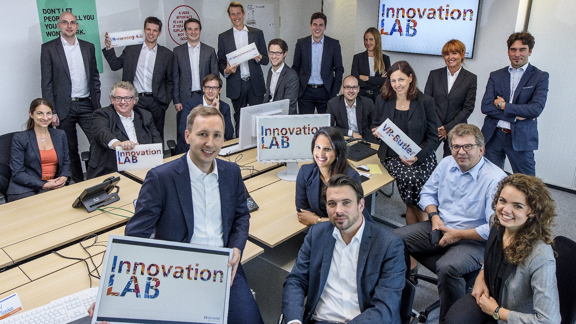 The image shows a group of professionally dressed people in an office setting. Some are holding signs that say 'Innovation LAB'. The environment looks like a workspace or meeting room, and there are computer monitors and a screen displaying 'Innovation LAB' in the background. The group appears to be participating in a collaborative or innovative event or workshop.