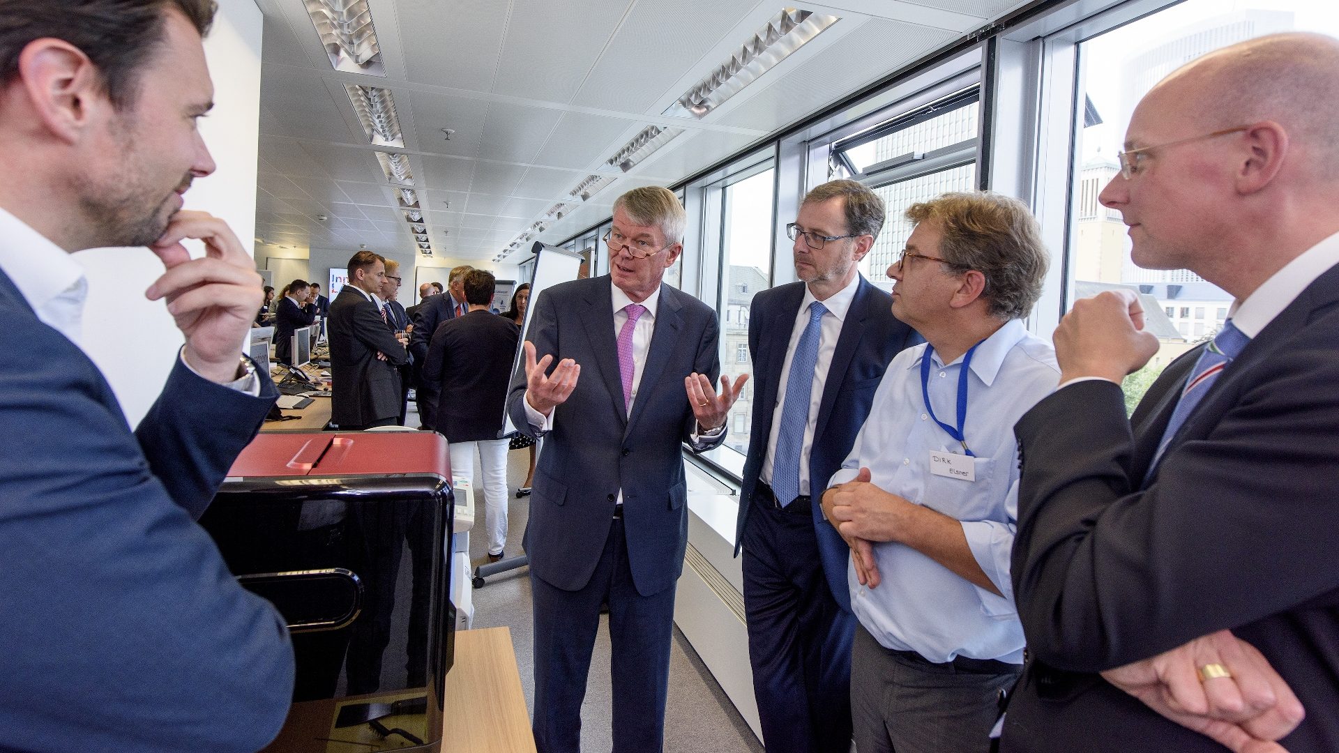 The image shows a group of five men engaged in a conversation in an office or conference setting. They are dressed in business attire, and the environment appears professional, with other people and office furniture visible in the background.
