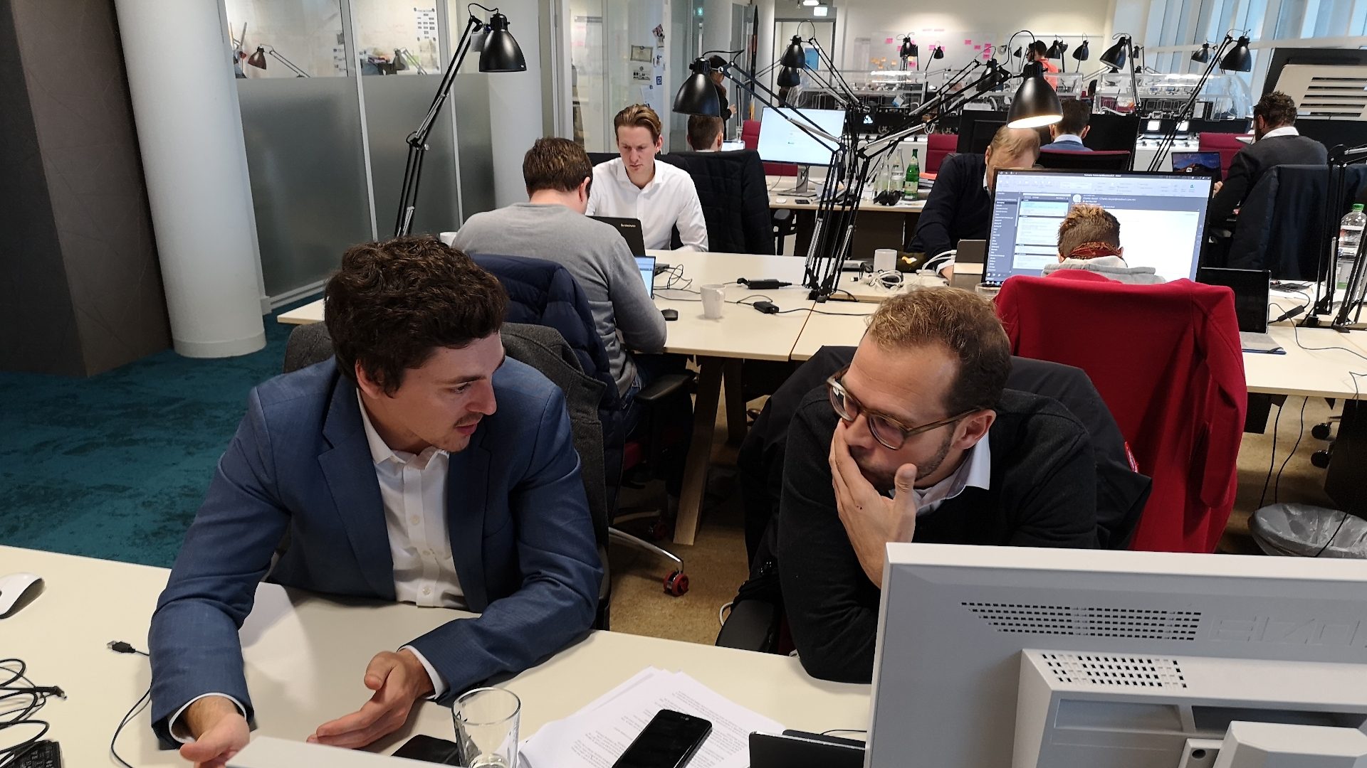 The image shows a busy office environment with several people working at desks. Two men are sitting in the foreground, engaged in conversation and looking at some papers or a computer screen. There are computers, paperwork, and other office equipment visible. Other people are also working at their desks in the background.