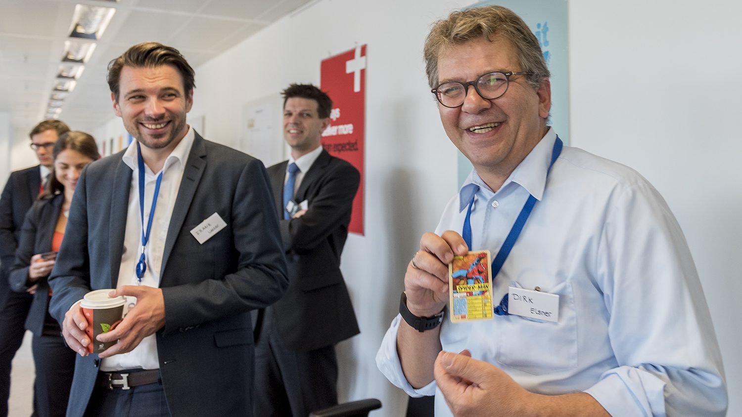 The image shows a group of people in a professional setting, likely an office. They are wearing formal clothing, some have lanyards with badges around their necks. One person is holding a colorful card and smiling, while another person is holding a coffee cup and also smiling. The atmosphere appears to be friendly and casual.