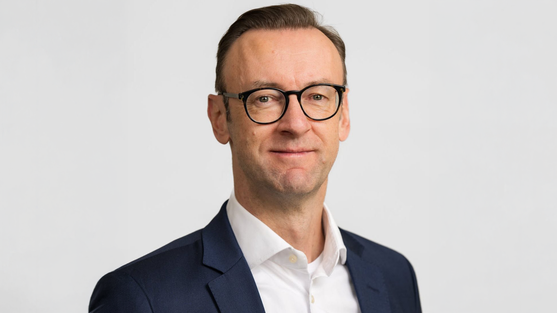 The image shows a man wearing glasses, a dark suit jacket, and a white shirt. He appears to be posing for a professional or formal portrait against a neutral background.