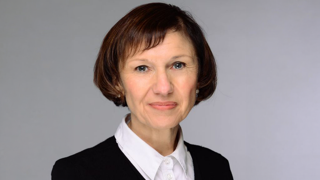 The image shows a woman with short, brown hair, wearing a white collared shirt and a dark jacket. She is facing the camera and smiling slightly, and the background is plain and light-colored.