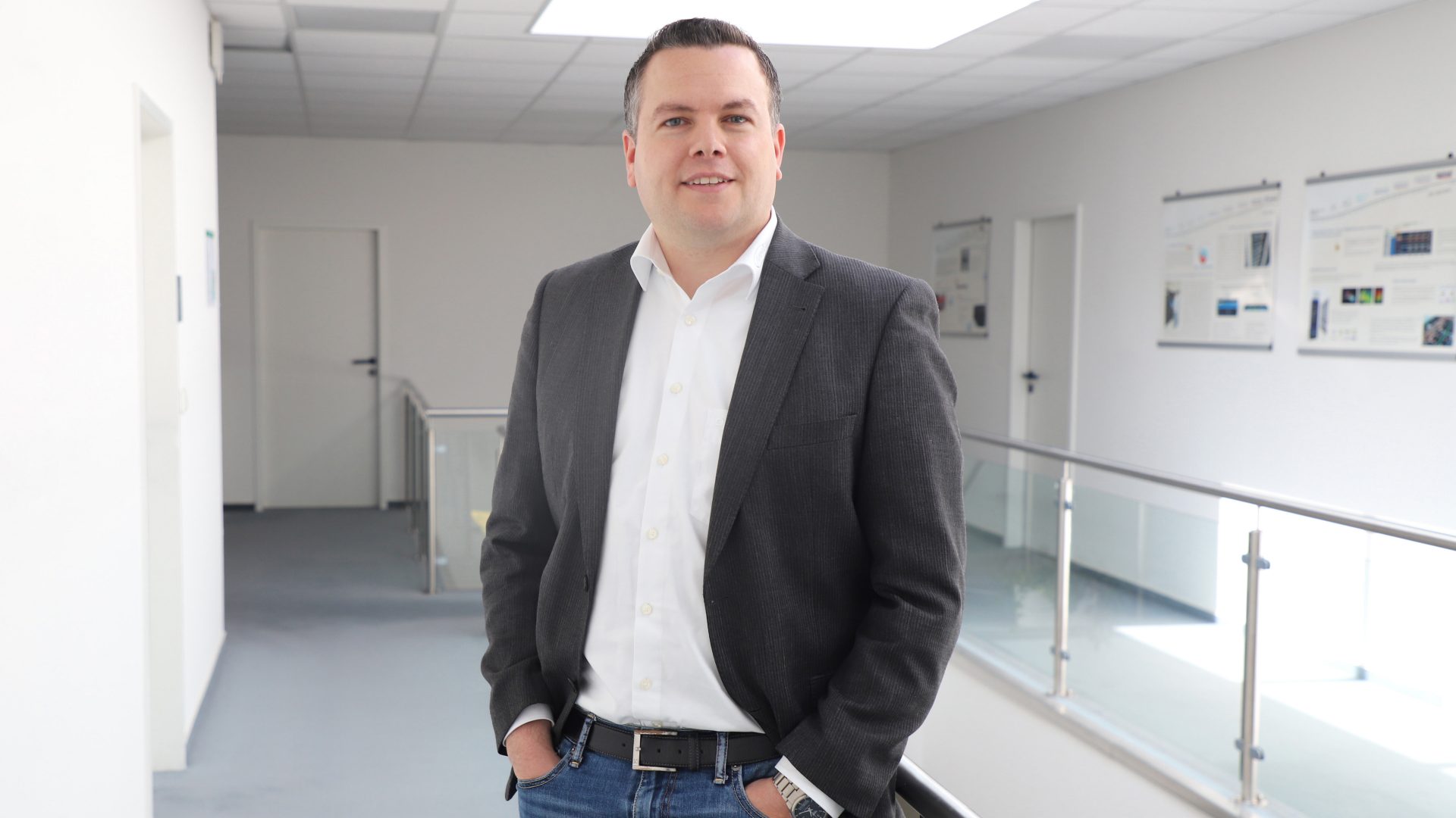 The image shows a man standing in a hallway or corridor of a modern building. He is wearing a dark blazer, white shirt, and jeans. The environment appears office-like, with posters or information boards on the wall and glass railings.