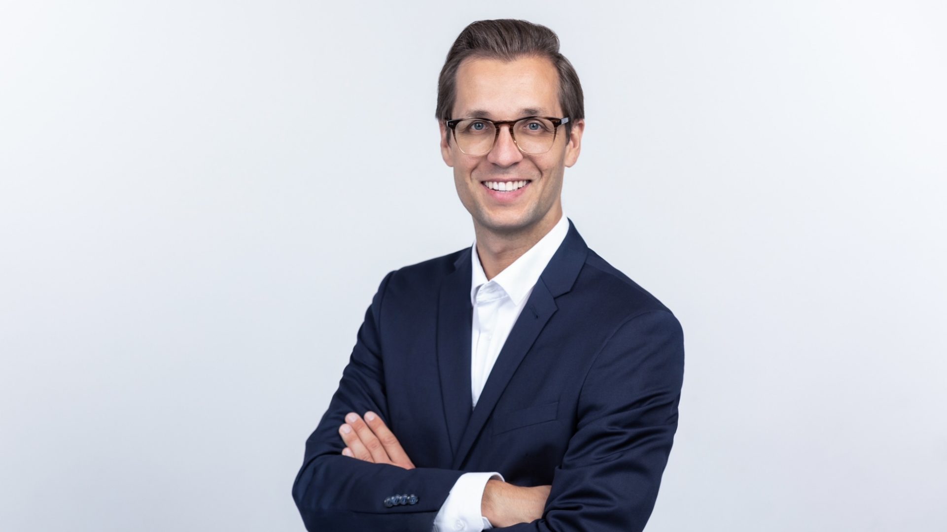 The image shows a man standing against a plain light background. He is wearing glasses, a dark suit, and a white shirt. He has his arms crossed and is smiling, projecting a professional and confident appearance.