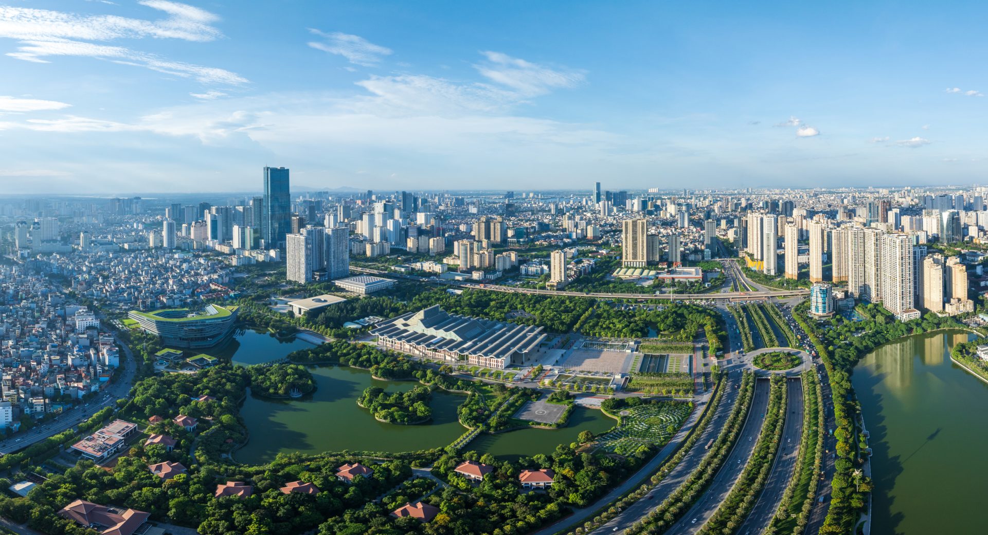 Hanoi Stadt The image depicts a panoramic view of a modern cityscape under a clear blue sky. Key features visible in this image include:
1. **Skyscrapers**: Several modern high-rise buildings dominate the city’s skyline, showcasing architectural advancement.
2. **Natural Landscape**: There are lush green areas, possibly parks or gardens, that provide a natural contrast to the urban environment.
3. **Water Bodies**: The presence of water bodies, likely lakes or rivers, that add to the scenic beauty and enhance the ecological balance of the city.
4. **Infrastructure**: Well-developed roads and pathways can be seen, indicating efficient urban planning and connectivity.
5. **Overall Ambiance**: The image presents an aerial view that encapsulates the harmonious blend of nature and modernity, reflecting a vibrant and well-structured metropolis.