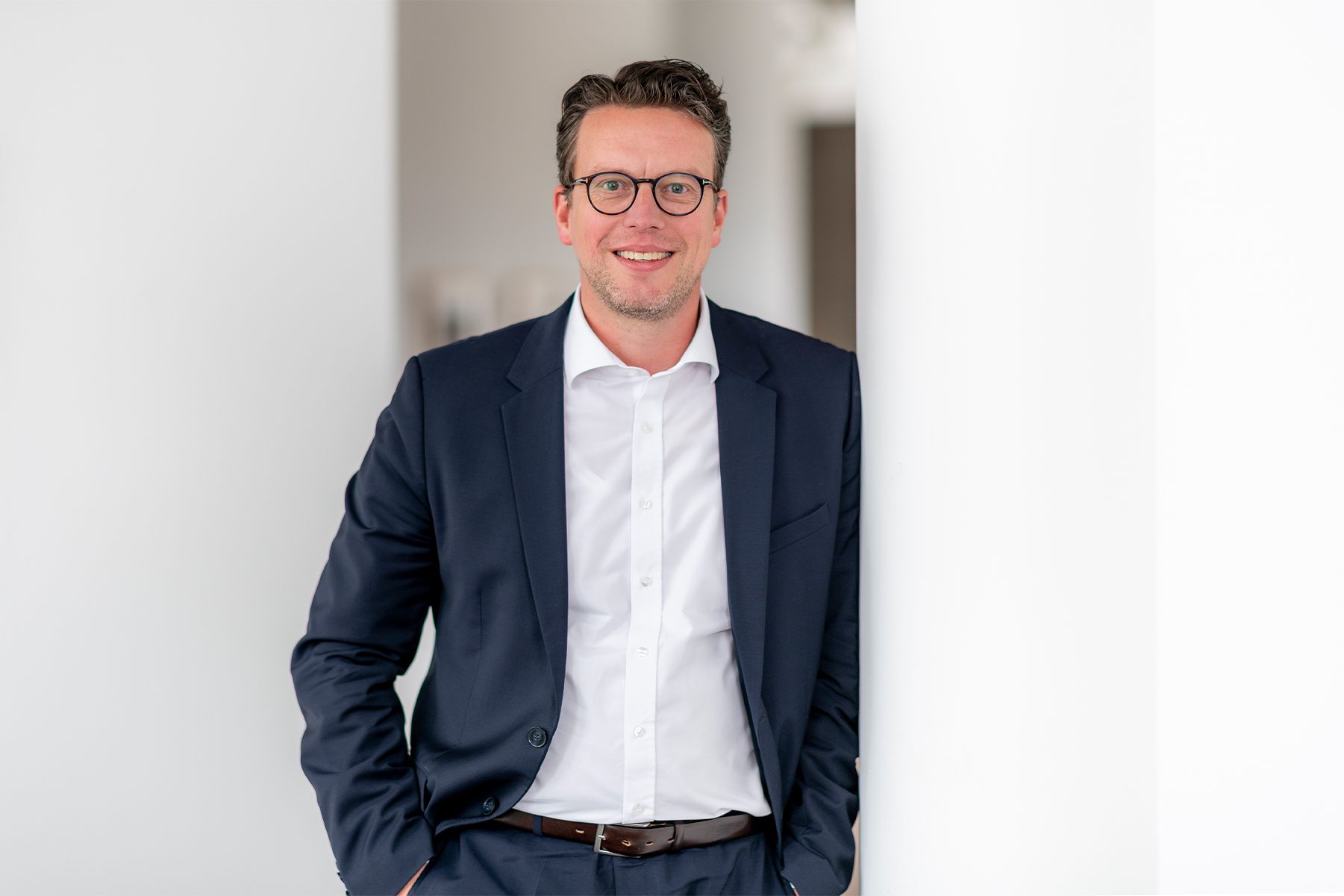 A man wearing a navy blue suit with a white shirt, standing against a white wall. His hands are in his pockets.