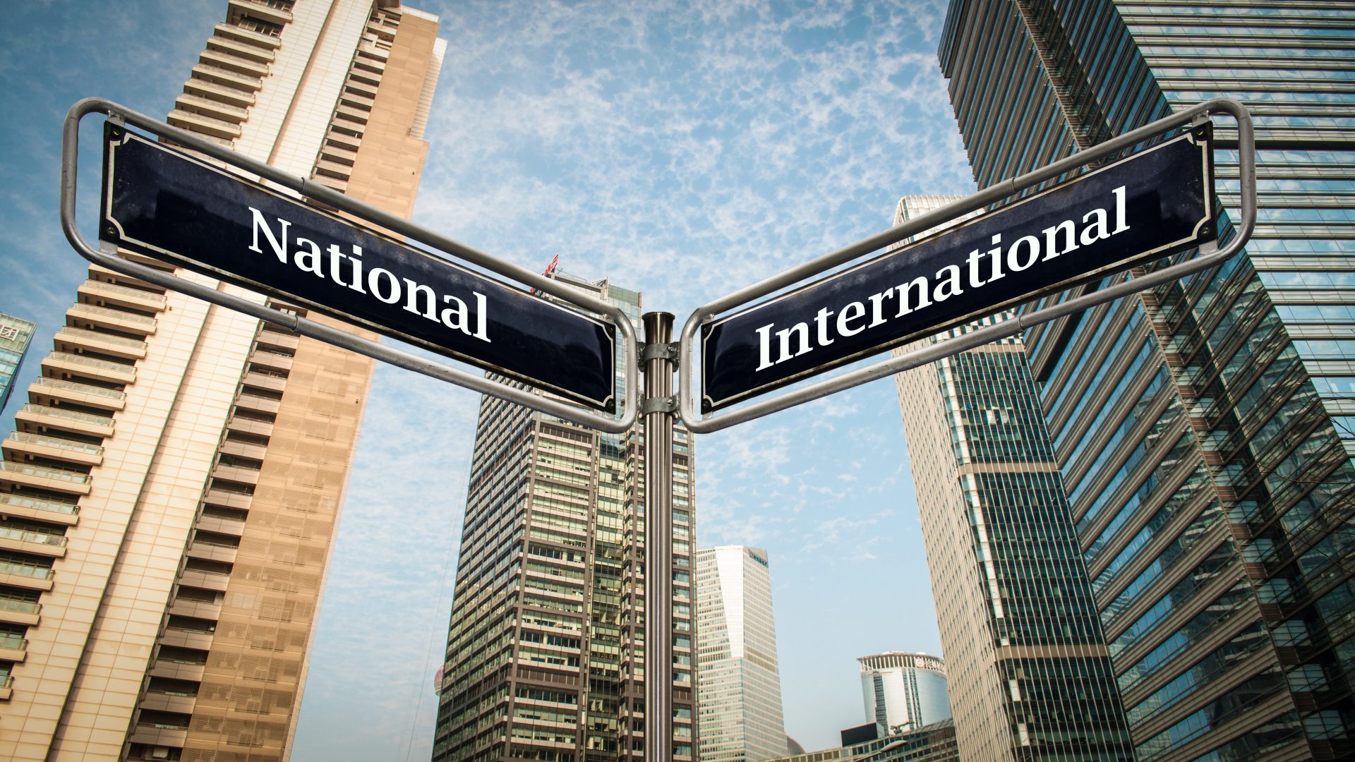 The image depicts a cityscape setting with tall modern buildings under a clear sky. Prominently featured in the foreground are two street signs, one labeled "National" and the other "International," suggesting a metaphorical crossroads or choice between local and global matters or directions.