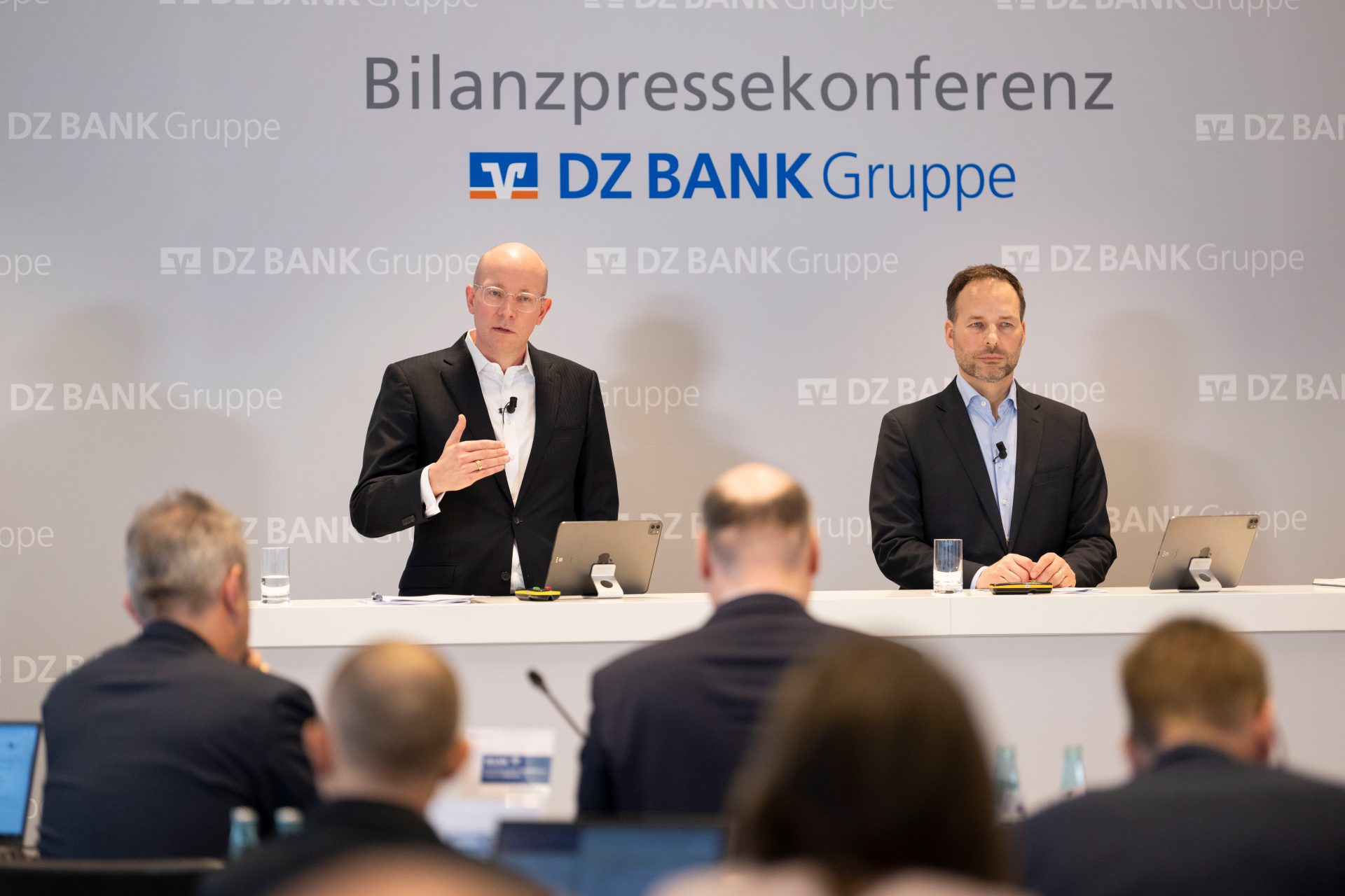 The image shows a corporate conference setting with a presentation or press conference backdrop featuring the DZ BANK Gruppe logo and the text "Bilanzpressekonferenz." The scene includes individuals sitting and listening, likely attending a business meeting or financial report presentation.