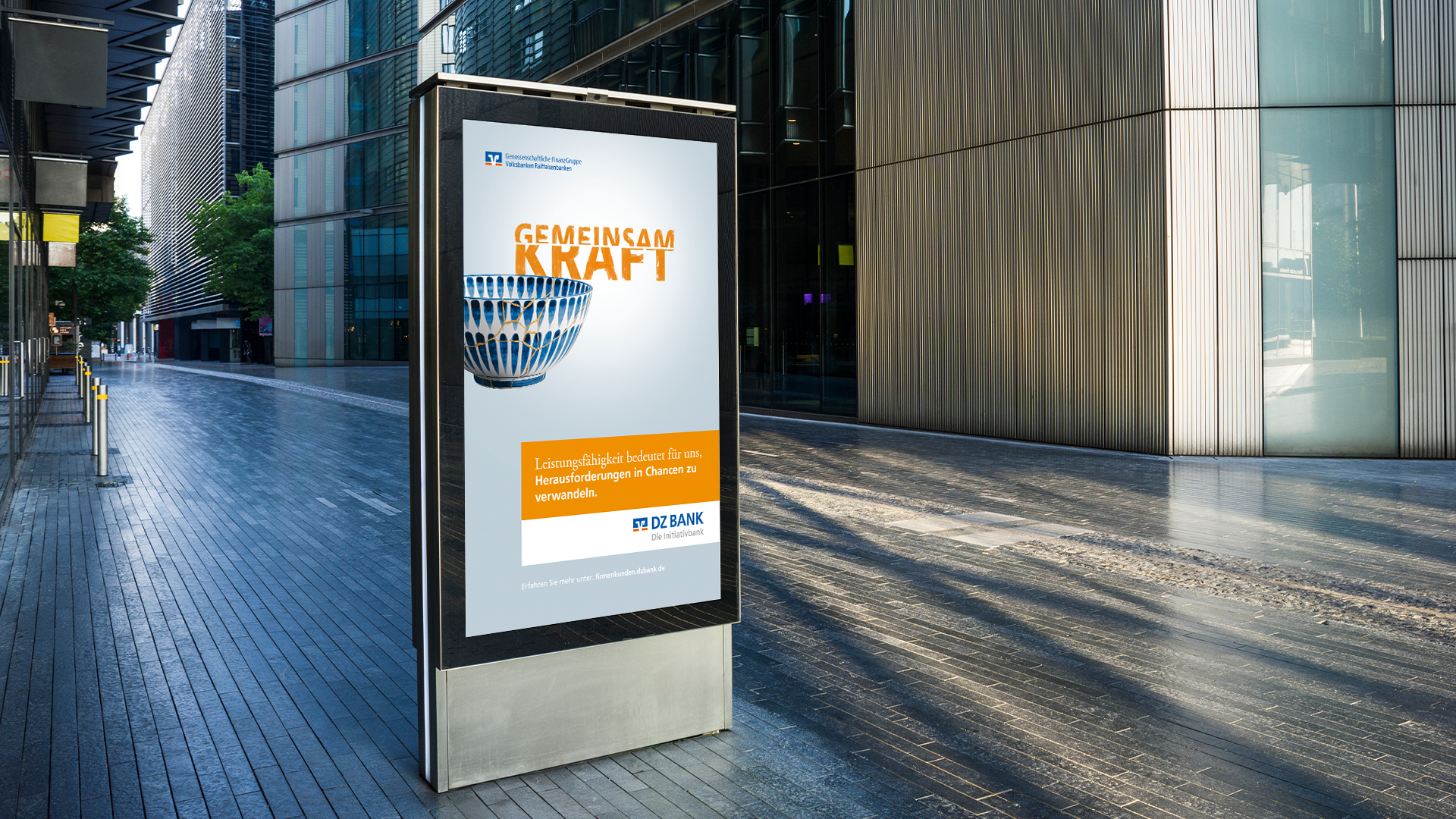 The image shows a sleek urban setting with tall modern buildings lining a pedestrian pathway. In the foreground, there is a large advertising billboard displaying promotional content.
**Details of the Image:**
1. **Billboard Advertisement**:
- **Text**: The advertisement prominently features the words "GEMEINSAM KRAFT," which translates to "TOGETHER POWER" in English.
- **Graphics**: Beneath the text is an image of a globe, symbolizing unity and collaboration.
- **Additional Text**: Underneath the main message, there’s more text in a white box on an orange background containing details or a slogan, likely promoting a message of strength or unity.
- **Logo or Branding**: There are logos or indicators of a company or organization at the top and bottom of the ad.
2. **Setting**:
- **Architecture**: Modern design with glass facades and clean lines.
- **Lighting**: It appears to be daytime, creating a reflective environment with shadows suggesting sunshine.
3. **Color Scheme**:
- **Prominent Colors**: Orange and white, possibly chosen for their eye-catching and positive significance.
- **Background**: Earthy and metallic tones in the architectural surroundings, complementing the advertisement's bright colors.
This scene likely represents a message or campaign encouraging strength, cooperation, or community among people, organizations, or nations.