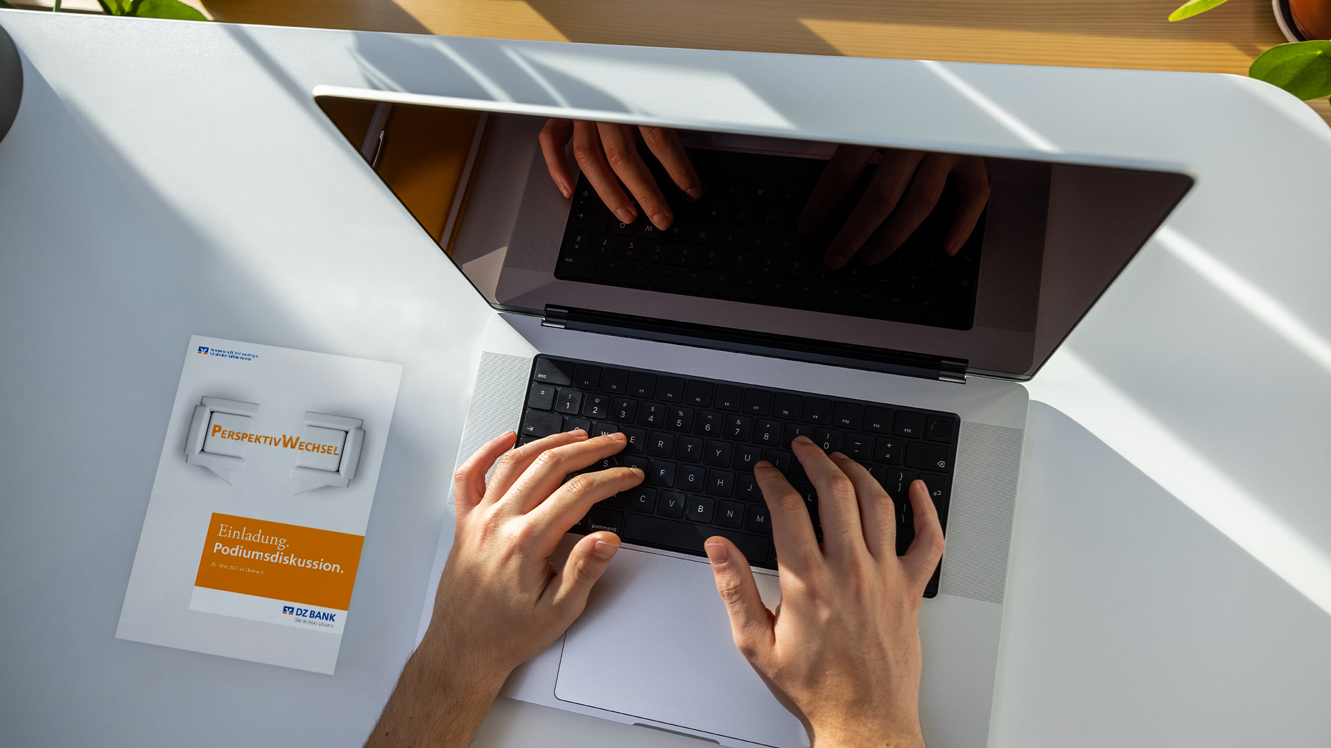 The image shows a person's hands typing on a laptop keyboard. The laptop is placed on a white desk. Next to the laptop is a binder titled "Finanzwesen" which suggests it could contain financial or business-related information or documents. The room is lit with natural sunlight, as evident from the shadow and sunlight reflections on the desk. There appears to be some greenery, likely from houseplants, in the background contributing to a bright and refreshing workspace atmosphere.