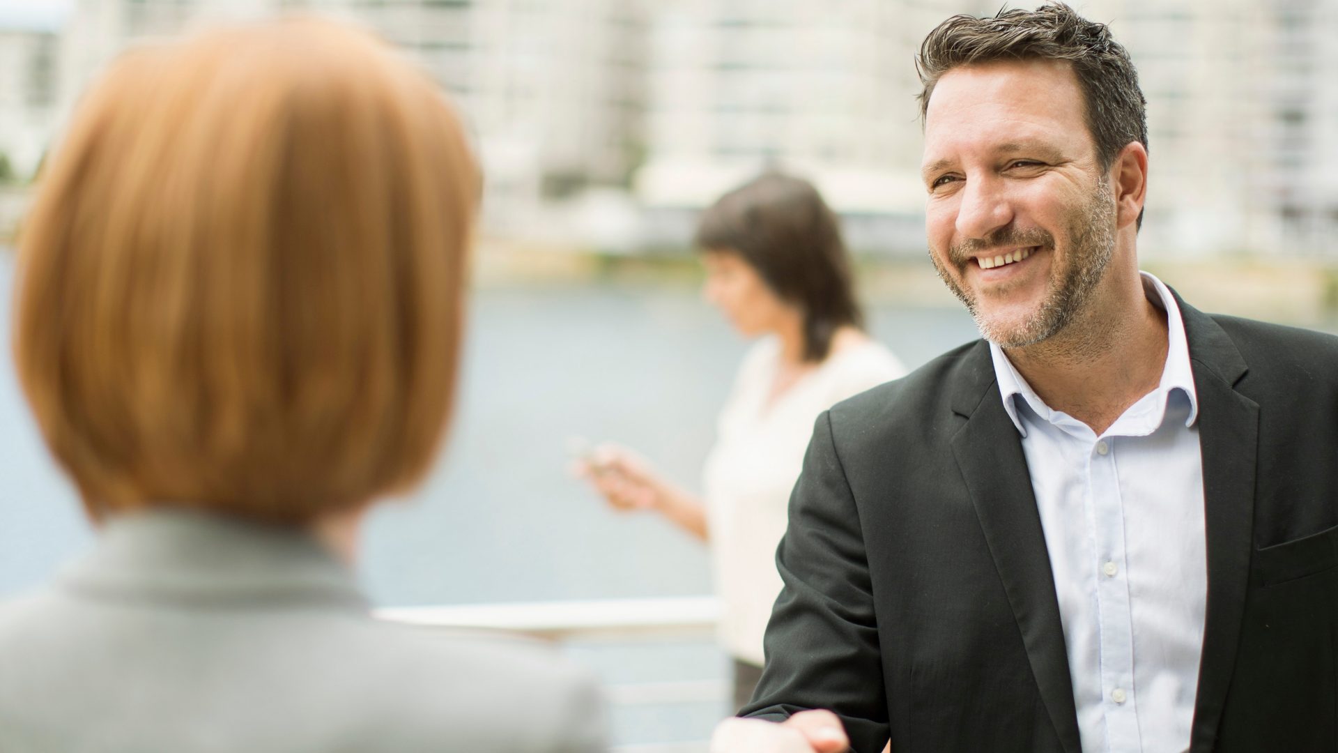Two people are interacting in an outdoor setting, possibly conducting business or having a conversation. The background includes blurred outlines of buildings or structures and another person is visible in the distance using a smartphone.