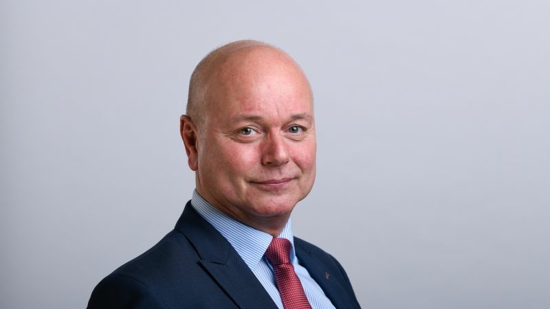 The image shows a person wearing a dark suit with a light blue dress shirt and a red tie. The background is plain and neutral.