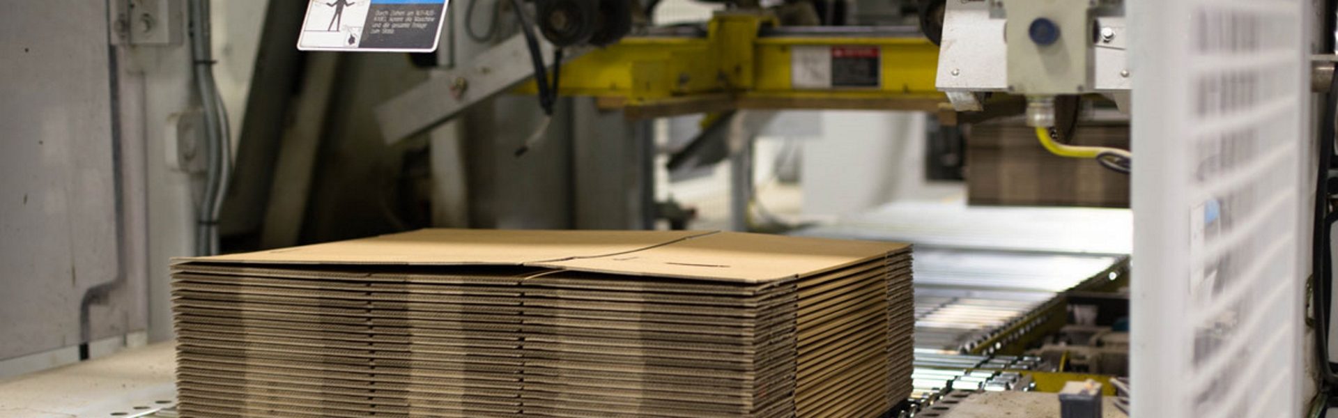 The image depicts a stack of cardboard boxes or sheets positioned under industrial machinery in a manufacturing setting. The machinery is primarily used for cutting, folding, or processing cardboard materials, potentially for producing packaging or other purposes. The environment suggests automation or mechanized operations with visible mechanical components and control panels.