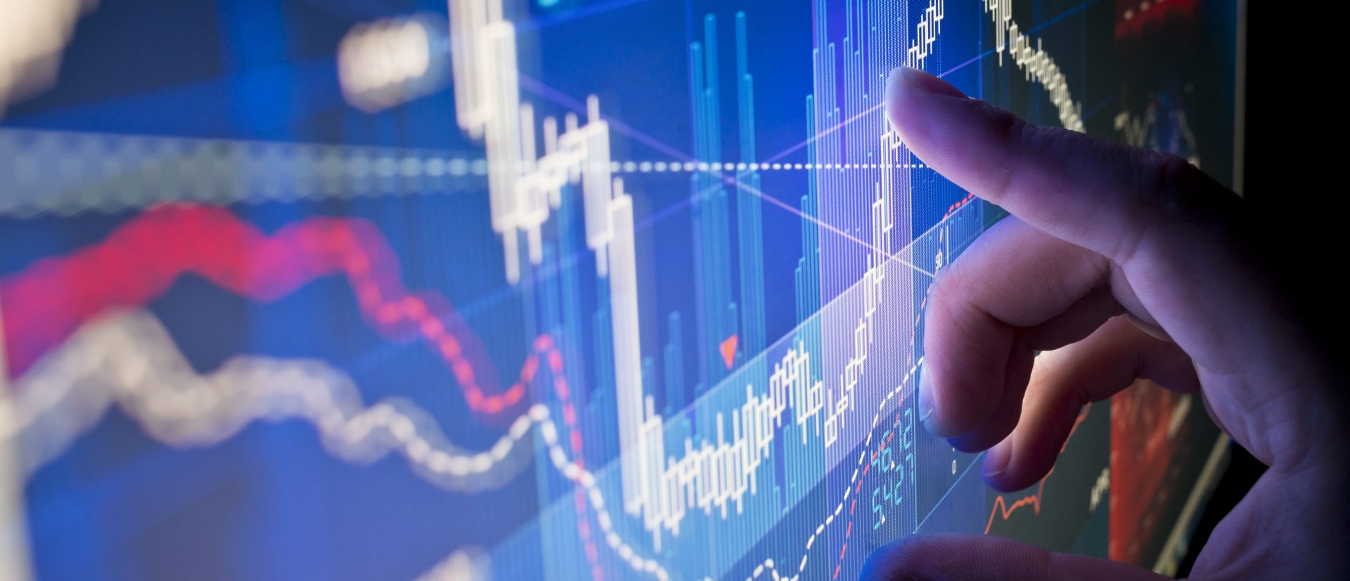 The image depicts a person's hand interacting with or pointing at a digital screen that displays financial data, likely related to stock markets or trading activities. The screen shows a variety of graphical representations such as line and bar charts that demonstrate fluctuations in data over time. The style and color arrangement suggests analysis of market trends, illustrating indices or levels that may be rising or falling. This type of visual is commonly used in financial advising, investment analysis, or stock market assessment contexts. The image conveys a sense of technology-driven financial decision-making process.