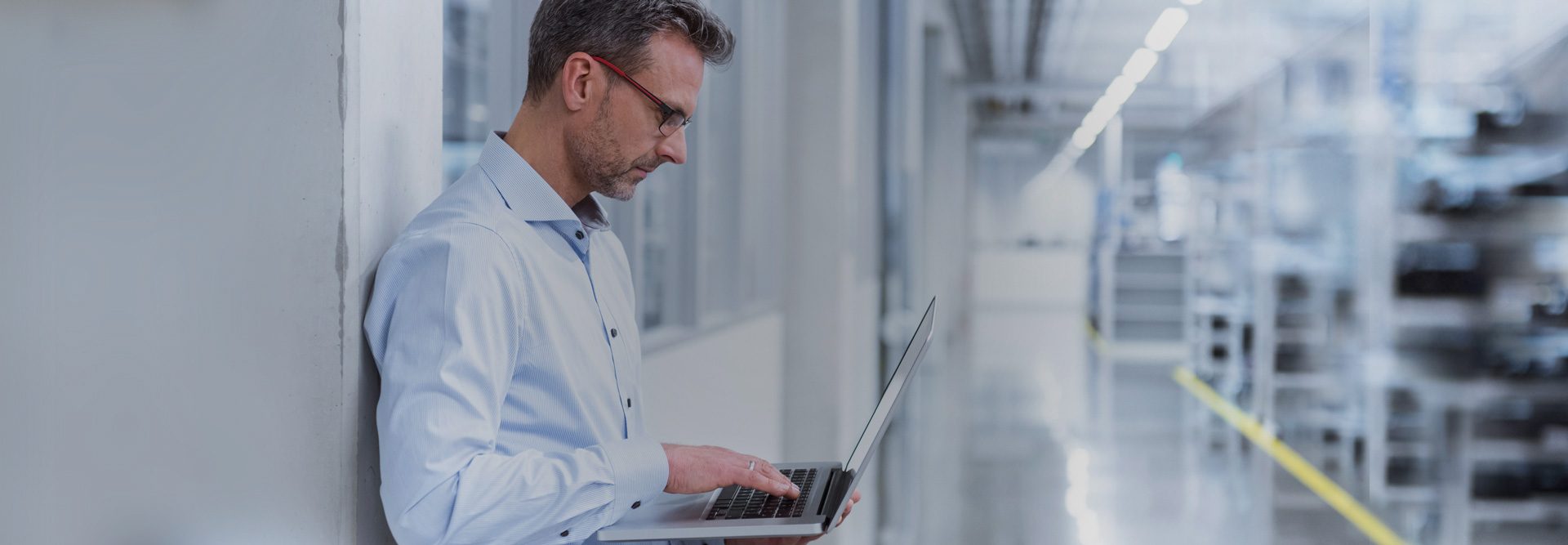 The image shows a person standing indoors holding a laptop in their hands. They are in what appears to be a modern, industrial or laboratory setting with fluorescent lighting and a shiny, reflective floor. There is a clear hallway or aisle that stretches to the background.